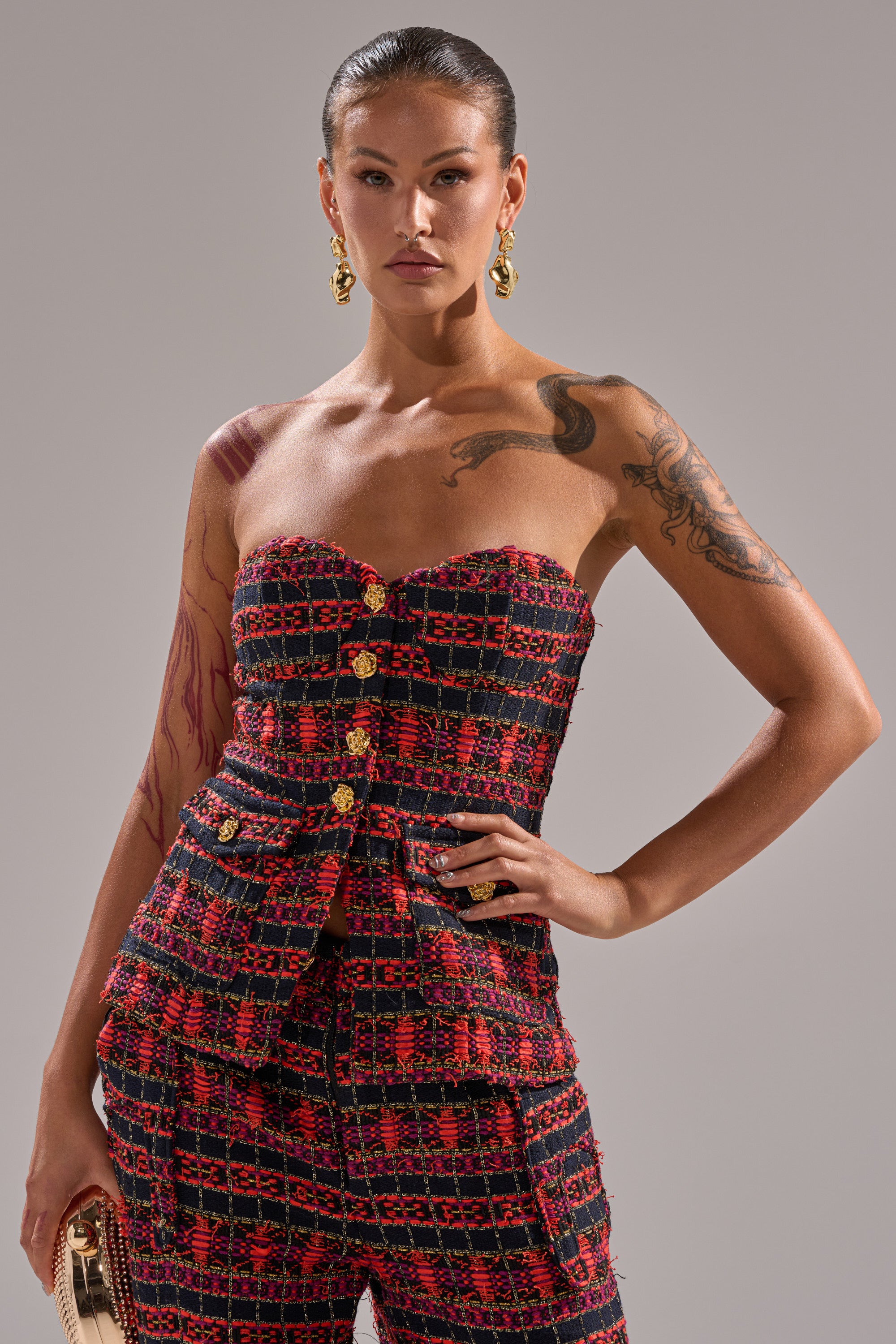 A woman poses confidently in the ONLY THINK ABOUT YOU CORSET IN TWEED, a strapless red and black plaid piece with gold buttons, styled with gold hoop earrings and a small gold clutch; she displays tattoos on her arm and shoulder. 0
