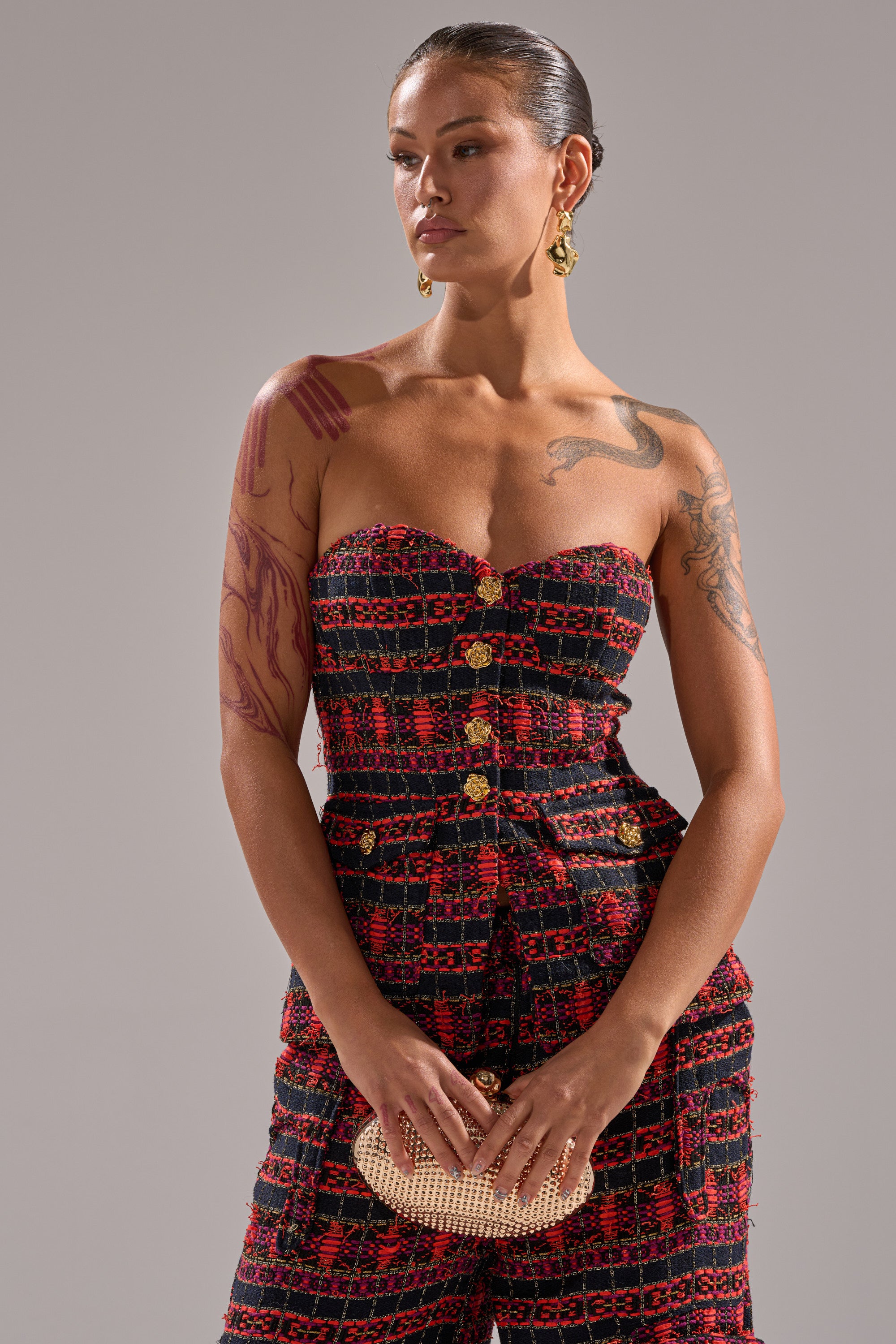A woman with tattoos wears the ONLY THINK ABOUT YOU CORSET IN TWEED, styled with a beige clutch and gold drop earrings, standing against a plain gray background. 1