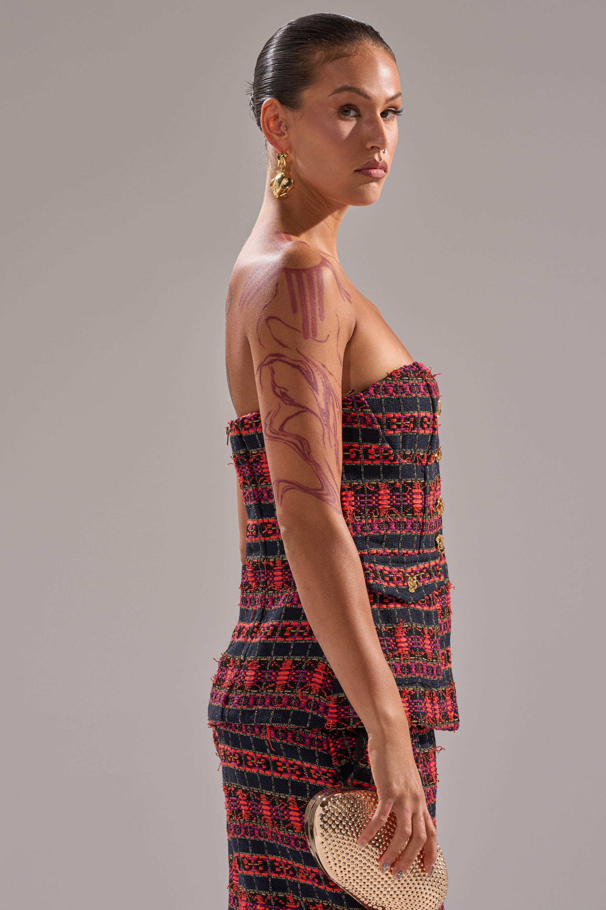 A woman with slicked-back hair wears a strapless red and black dress featuring the ONLY THINK ABOUT YOU CORSET IN TWEED, paired with gold earrings and a beige clutch. Bold purple-red body art adorns her arm as she stands against a plain background. 2