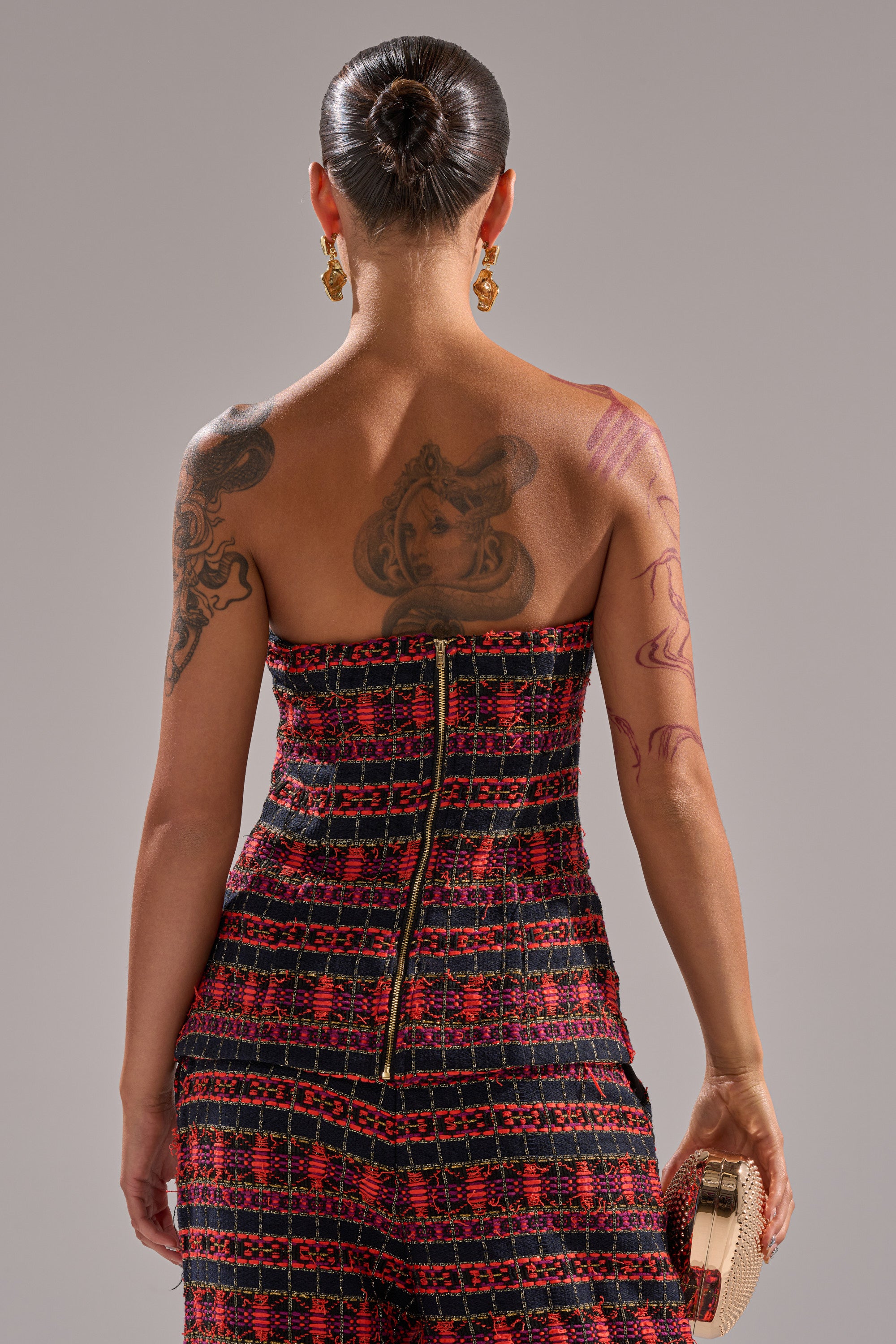 A woman faces away from the camera, wearing the ONLY THINK ABOUT YOU CORSET IN TWEED. She has a bun hairstyle, statement earrings, visible tattoos on her back and arms, and holds a small clutch in her right hand. 3