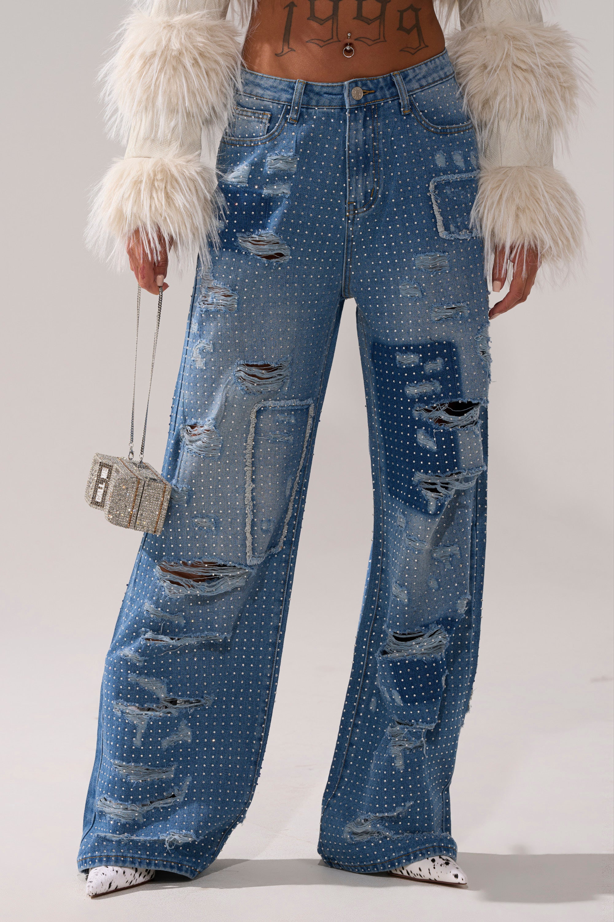 A person wears the LAY IT LOW RHINESTONE RELAXED DENIM, straight-leg blue jeans, paired with a furry white cropped jacket and holding a small sparkly handbag. Only their body is visible, not the face.