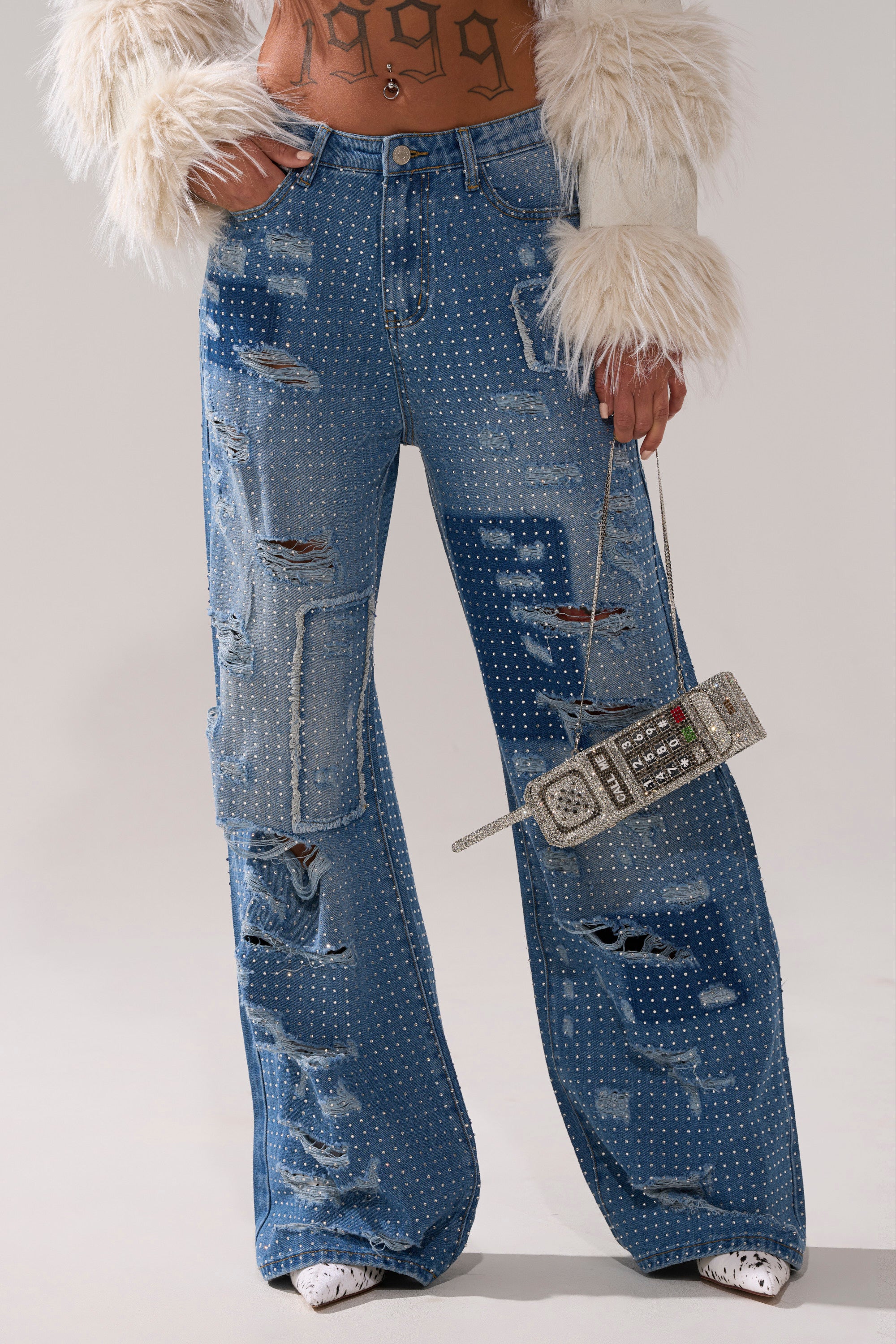 Wearing the LAY IT LOW RHINESTONE RELAXED DENIM and a furry white jacket, they hold a vintage cell phone-shaped purse, showing off a "1999" tattoo across their stomach.