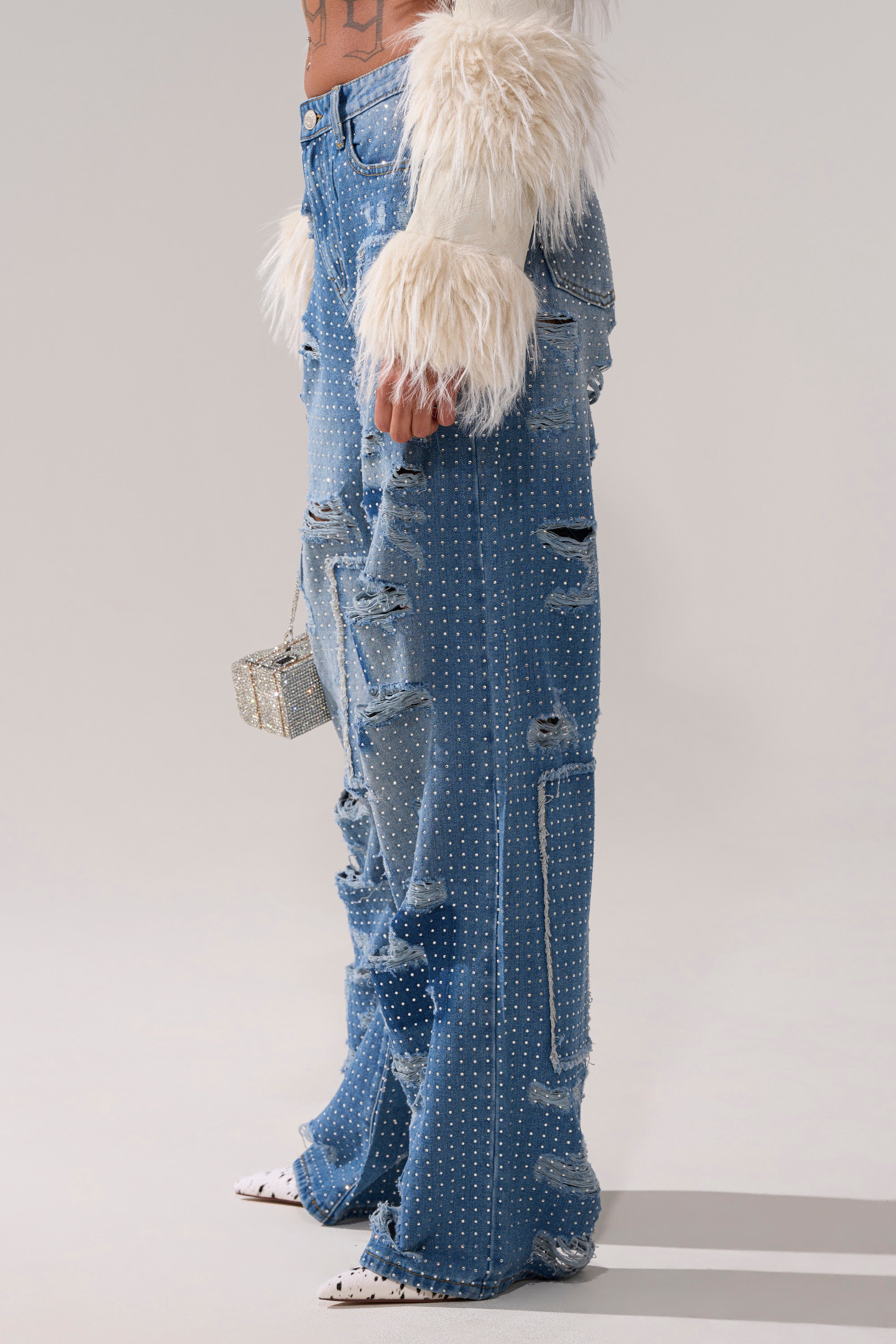 Wearing the LAY IT LOW RHINESTONE RELAXED DENIM, a furry white long-sleeve top, white pointed shoes, and holding a glittery small purse, the person stands against a plain background.