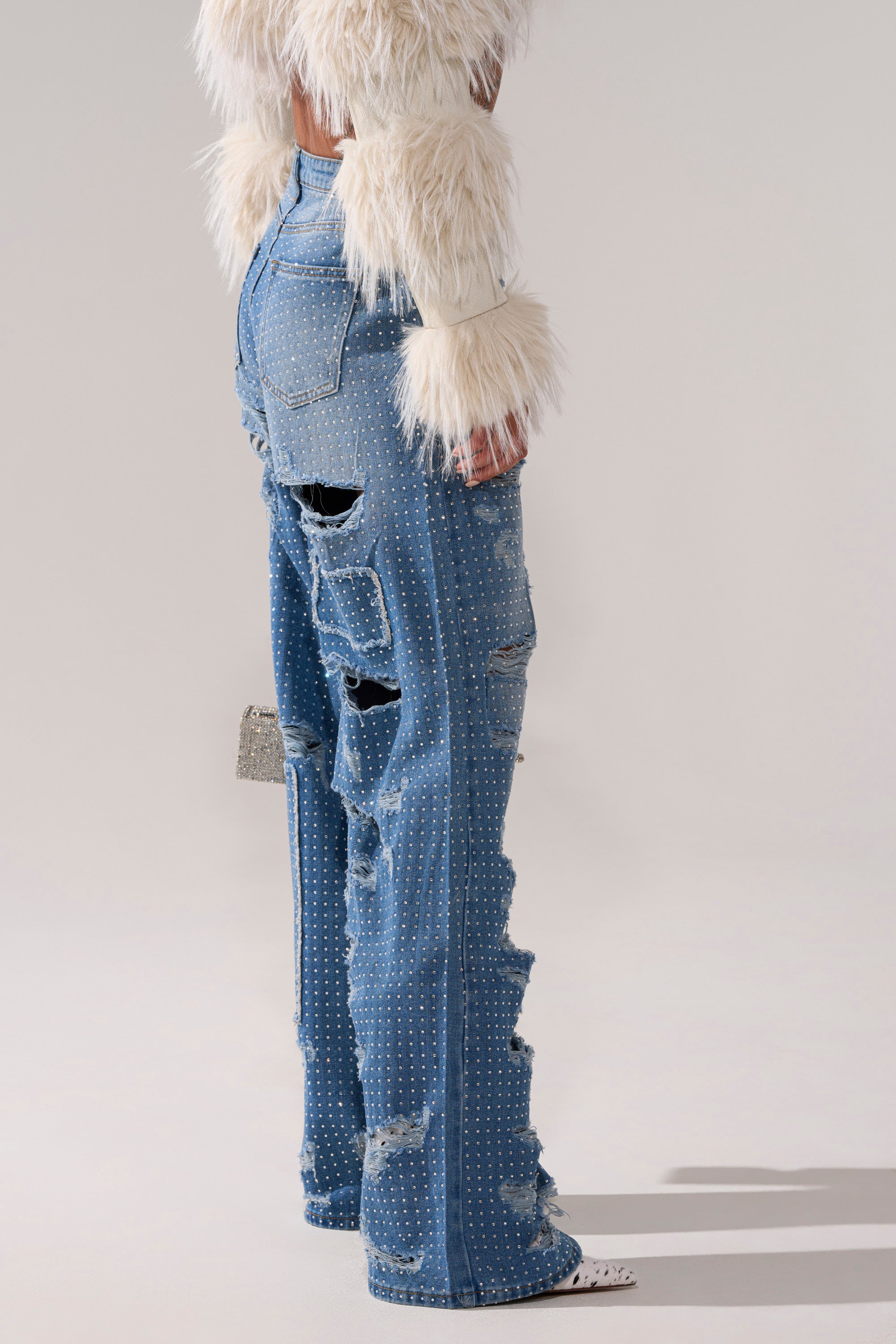 A person wears a white faux fur top and LAY IT LOW RHINESTONE RELAXED DENIM—high-rise, light blue distressed jeans with silver studs—against a neutral background.