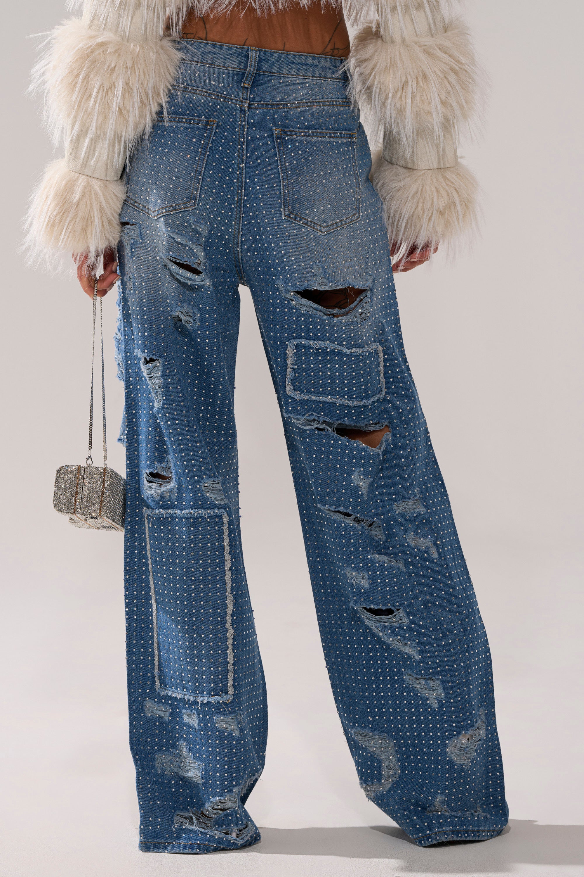 Wearing the LAY IT LOW RHINESTONE RELAXED DENIM, styled with a fluffy cream jacket and a glittery handbag, this look features distressed high-rise jeans with rhinestone details and a relaxed straight-leg fit for effortless sparkle.