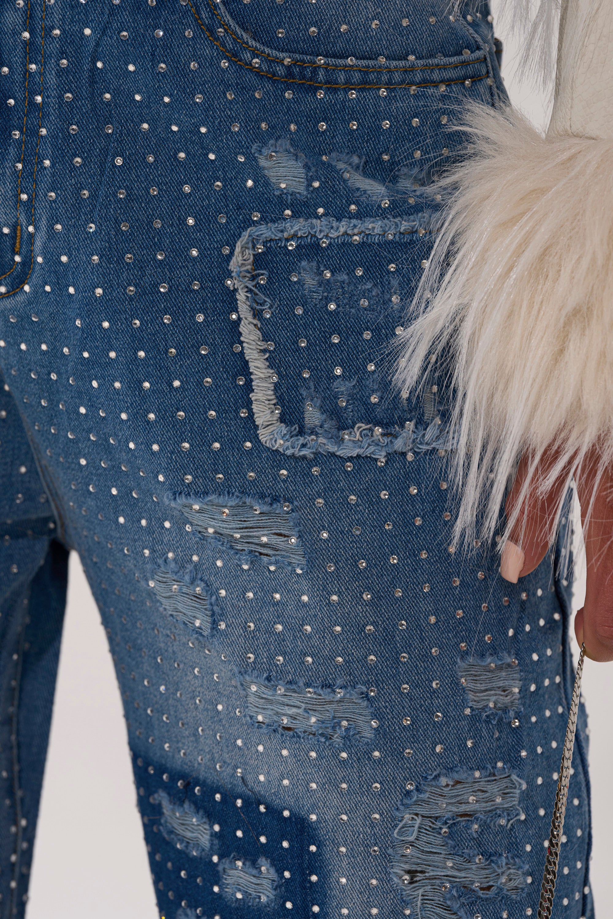 Close-up of a person wearing LAY IT LOW RHINESTONE RELAXED DENIM—distressed, high-rise jeans with silver studs and frayed edges, styled with a white furry sleeve as their hand rests casually by their side.