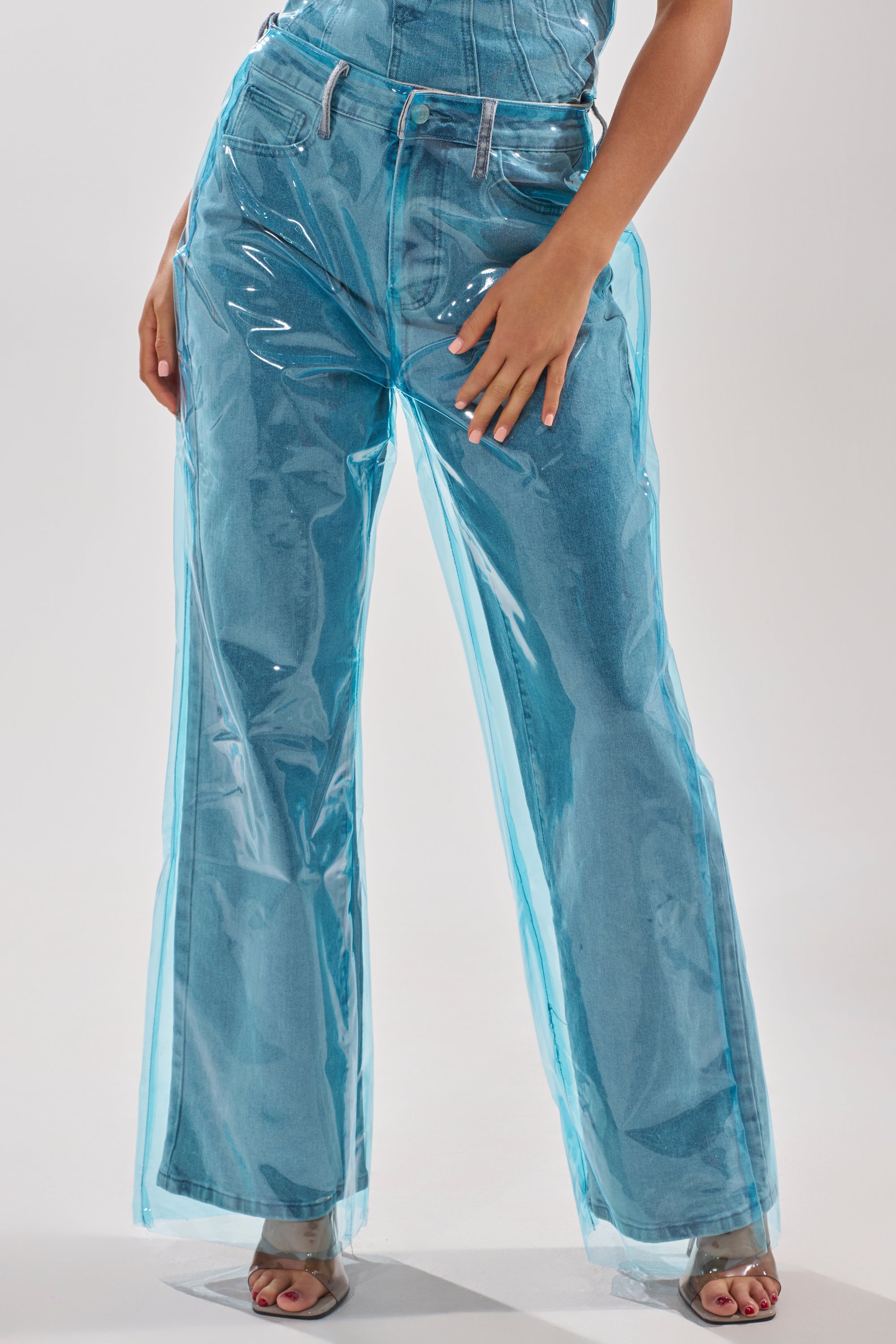 A person wearing ELECTRIC FEEL WIDE LEG DENIM featuring a shiny, transparent blue PVC overlay and open-toed heels, posing with one hand on their thigh. 2