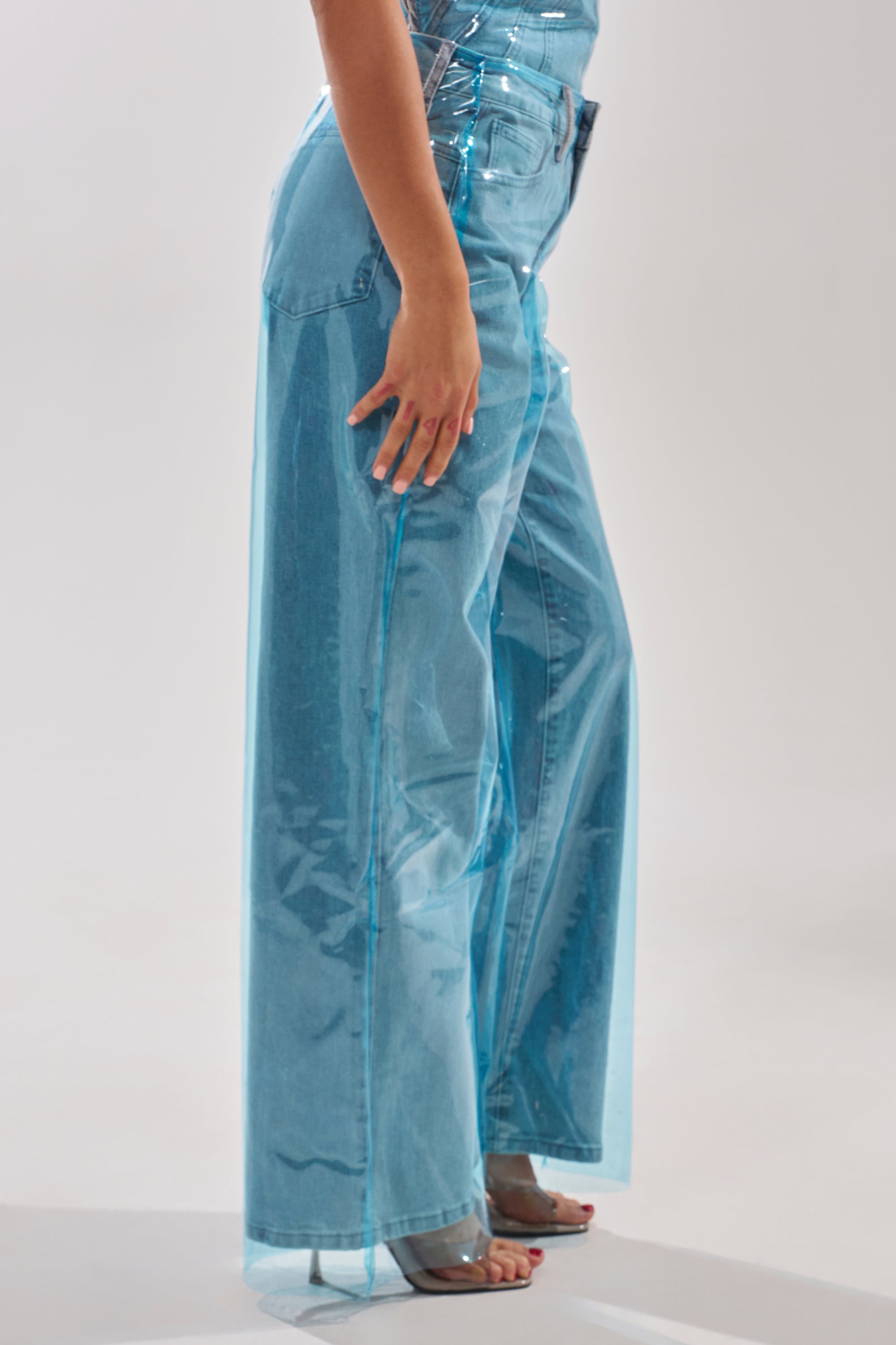 A person models the ELECTRIC FEEL WIDE LEG DENIM featuring a shiny blue PVC overlay. Only their lower torso, legs, and one hand are shown against a plain background. 3