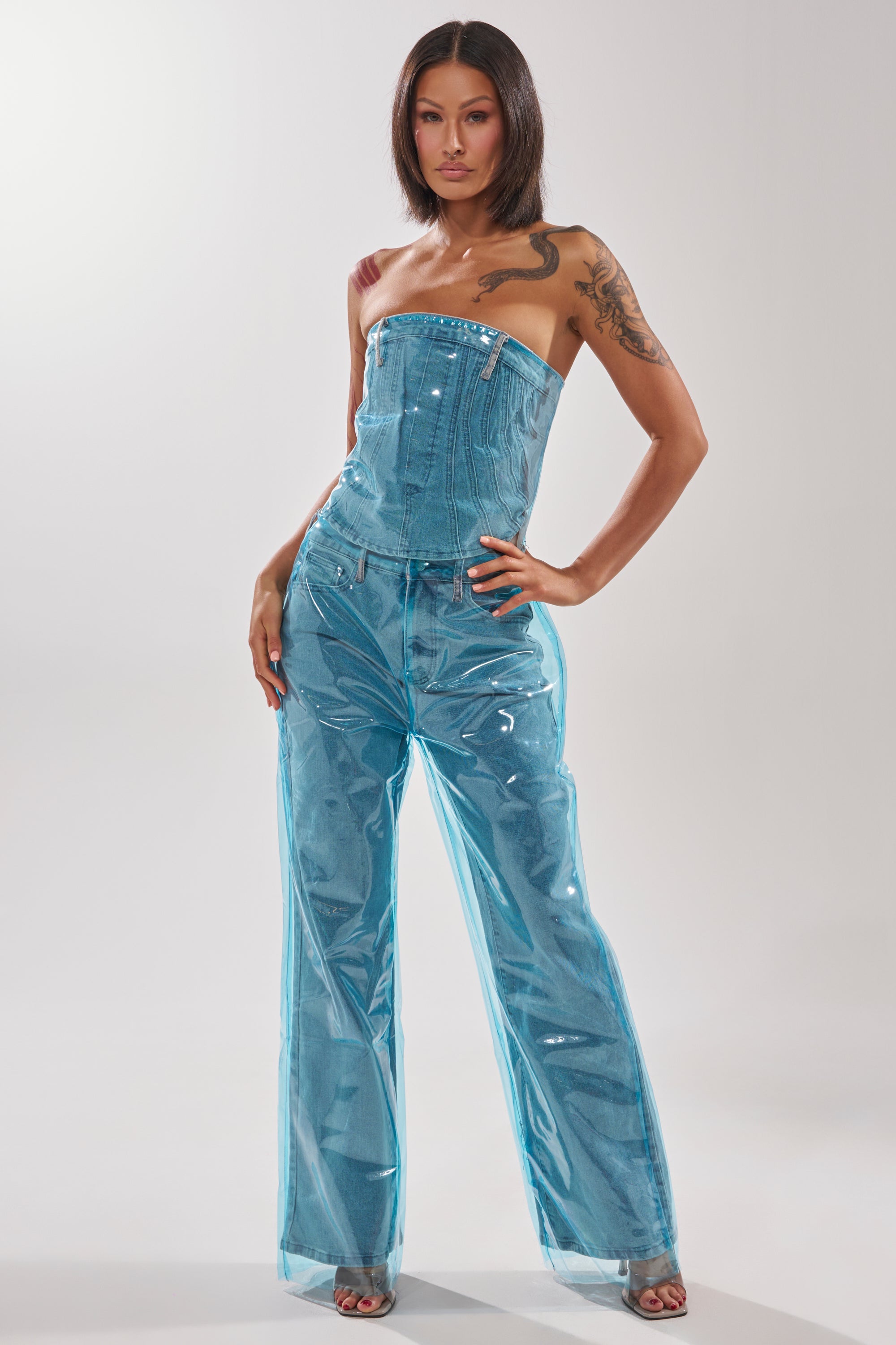 A woman models the ELECTRIC FEEL WIDE LEG DENIM with a strapless clear blue PVC overlay jumpsuit, revealing her arm tattoos against a plain light background. 5