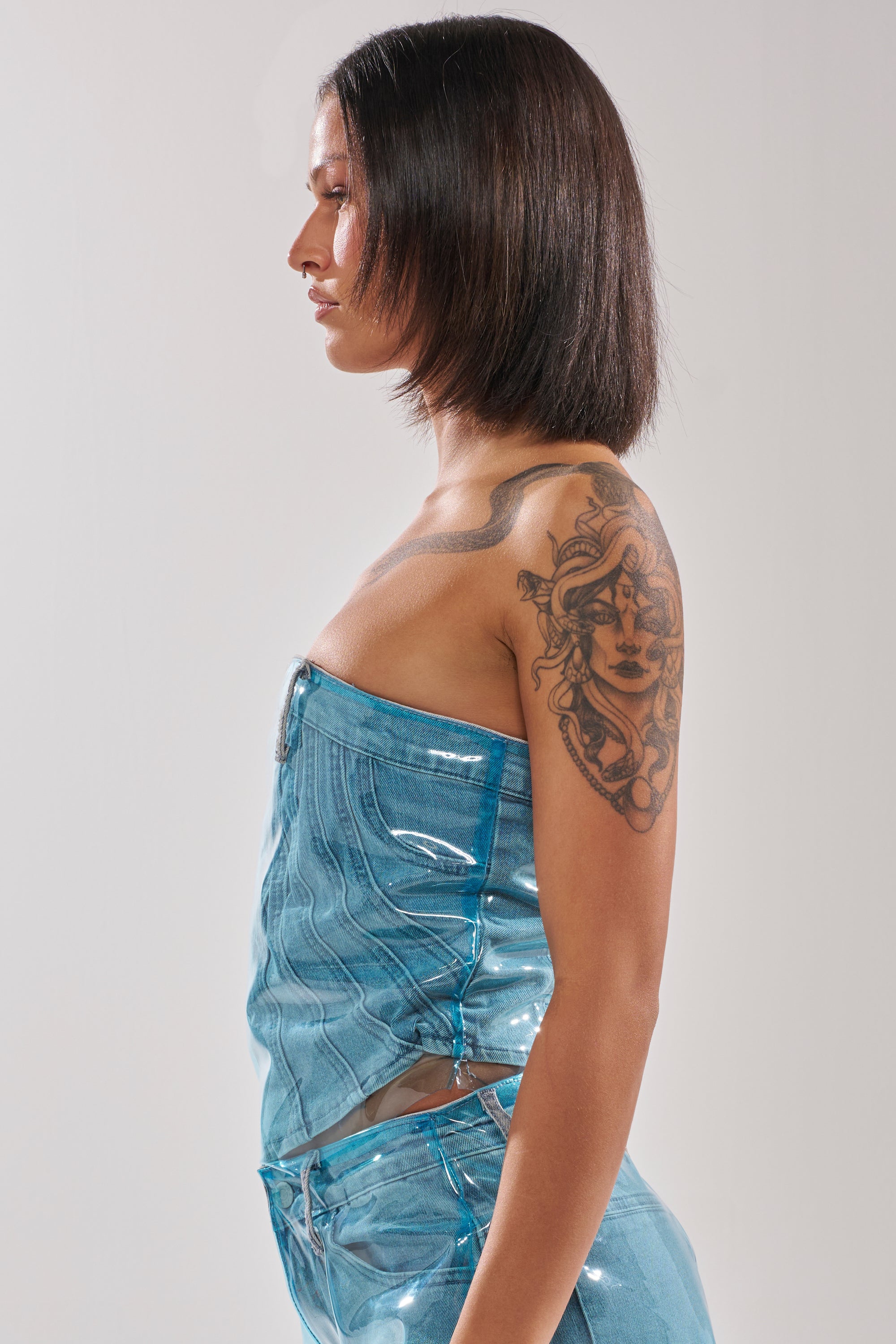 Woman with straight dark hair poses in profile, wearing the shiny ELECTRIC FEEL DENIM CORSET and matching PVC-overlay pants. She shows a large tattoo of a woman's face on her upper arm and back against a light plain background. 3