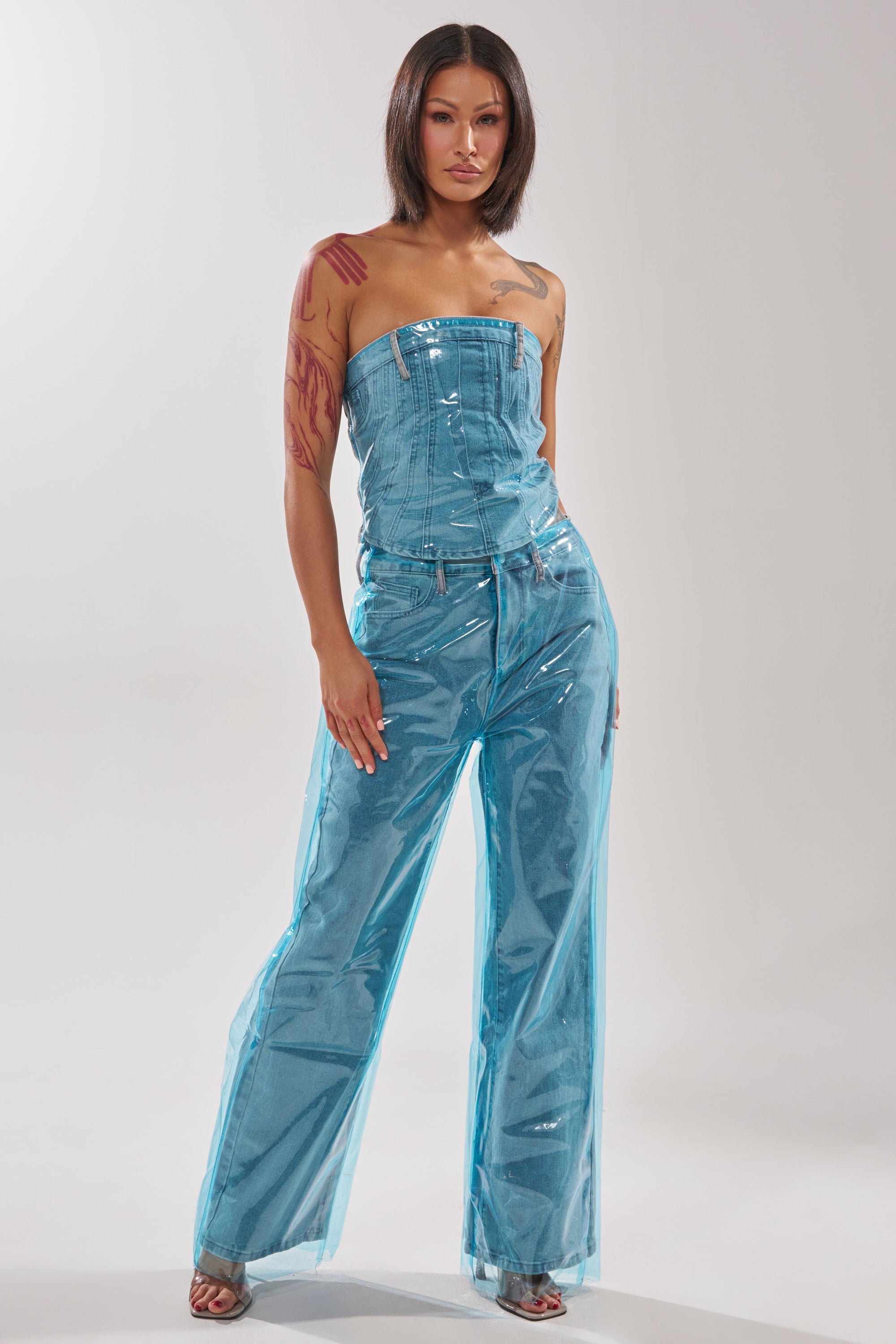 A woman with a bob haircut and visible right arm tattoos poses confidently in the ELECTRIC FEEL DENIM CORSET, a shiny strapless top styled with matching wide-leg pants that feature a plastic-like sheen. 5