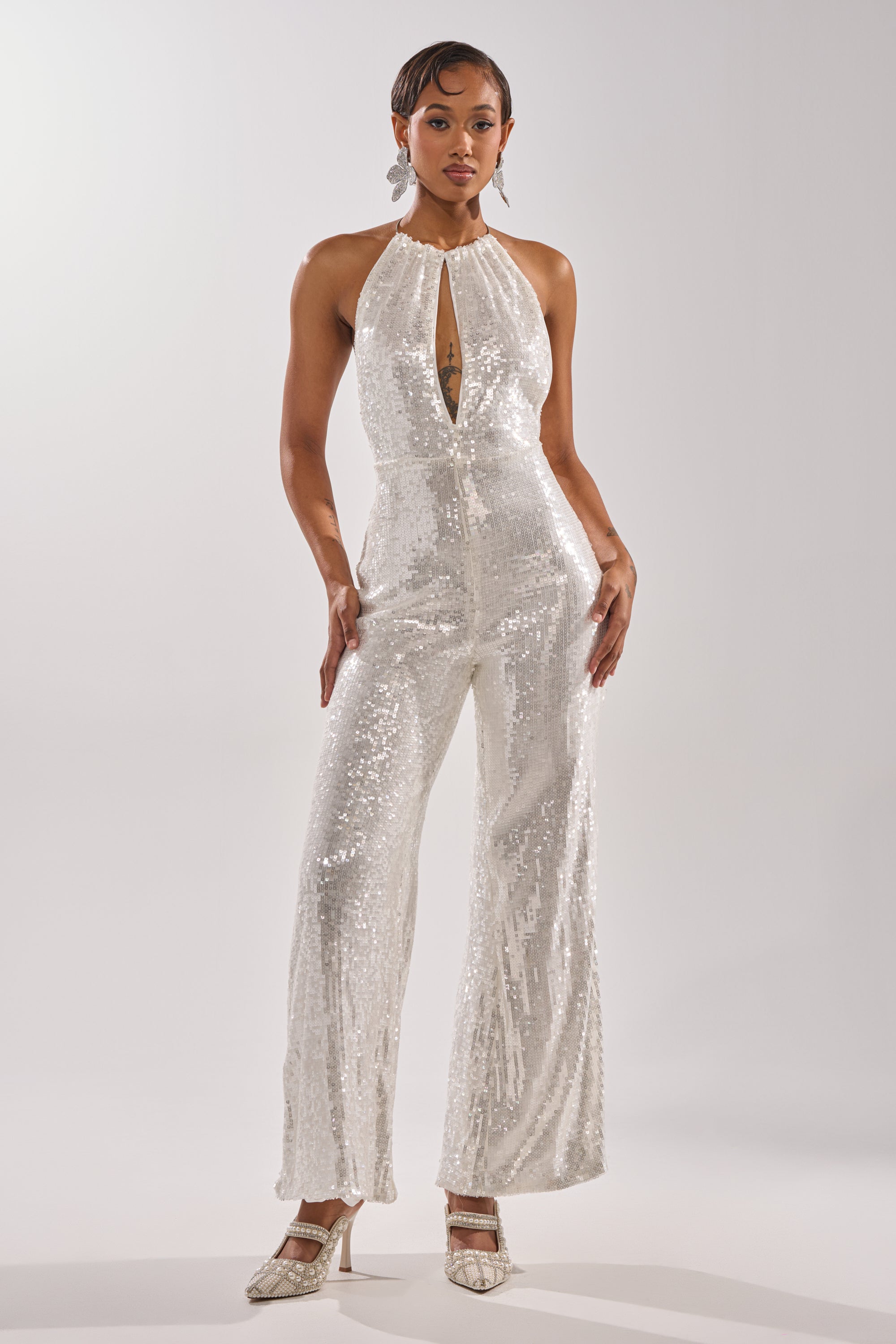 A woman models the ICE QUEEN SEQUIN JUMPSUIT, featuring a sleeveless halter neckline, front keyhole cutout, and shimmering white sequins. She accessorizes with sparkling earrings and pointed heels against a light background.