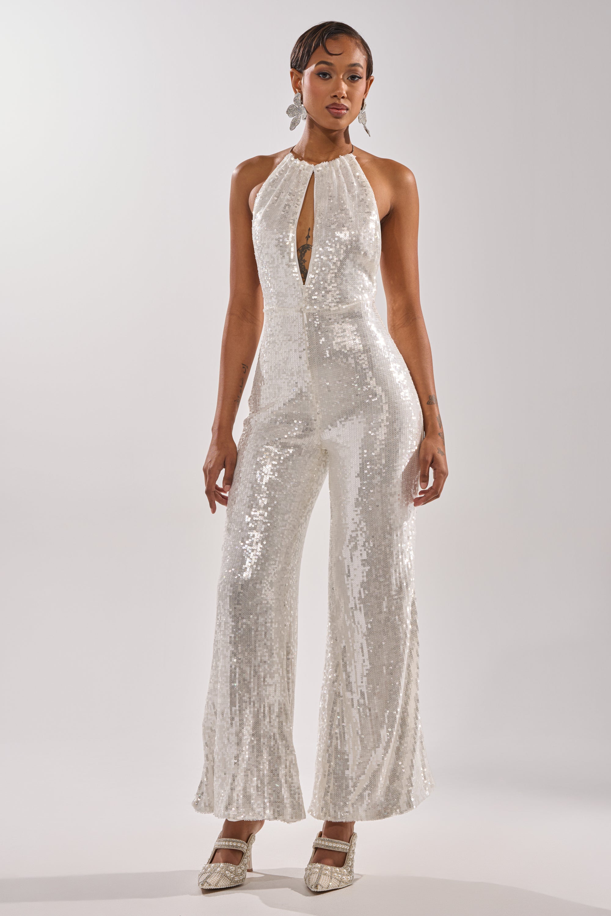 A woman poses against a plain background in the ICE QUEEN SEQUIN JUMPSUIT, a sleeveless white sequin piece with a halter neckline and flared legs. She completes the look with short hair, statement earrings, and pointed-toe heels.