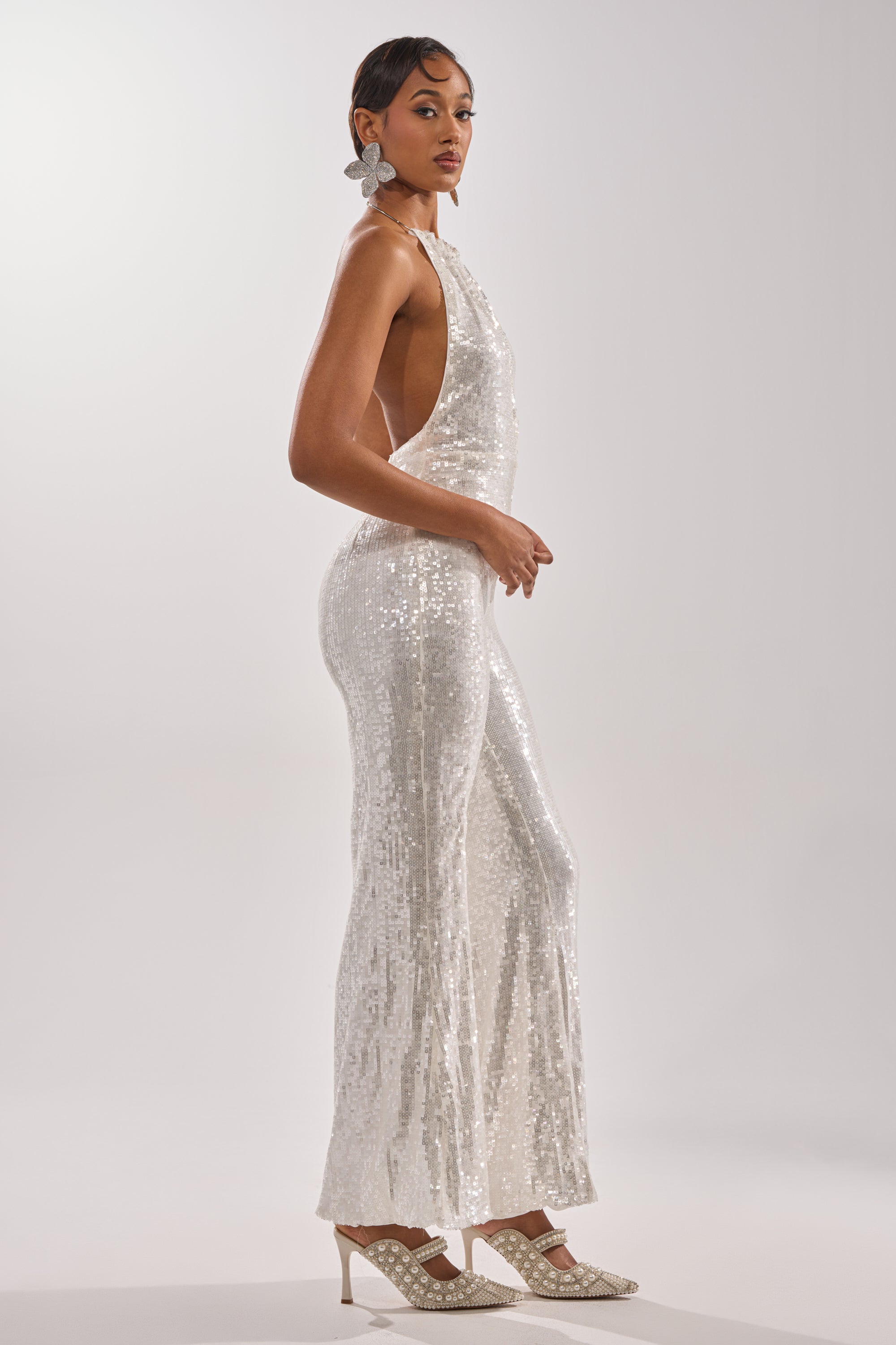 A woman with short hair and statement earrings models the ICE QUEEN SEQUIN JUMPSUIT—a sleeveless, backless, wide-leg style—paired with white heels against a plain, light background.