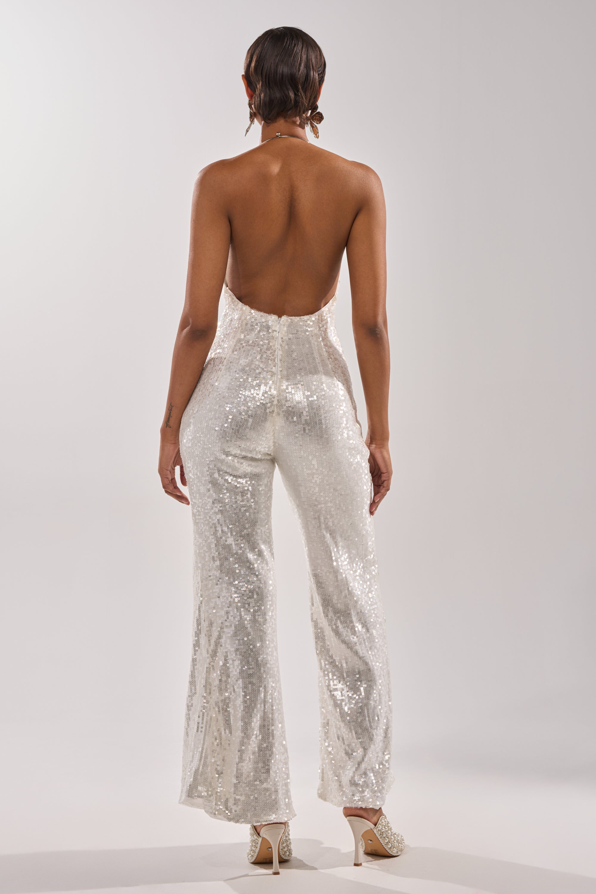 A person with short hair faces away, wearing the ICE QUEEN SEQUIN JUMPSUIT—a white, sleeveless, open-back design with a sparkling sequin finish and wide-leg silhouette—paired with high heels against a plain light background.