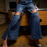 Wearing SPARKLE QUEEN WIDE-LEG DENIM and clear heels, a person poses by a pool table in a dimly lit room with wooden floors and brick walls.