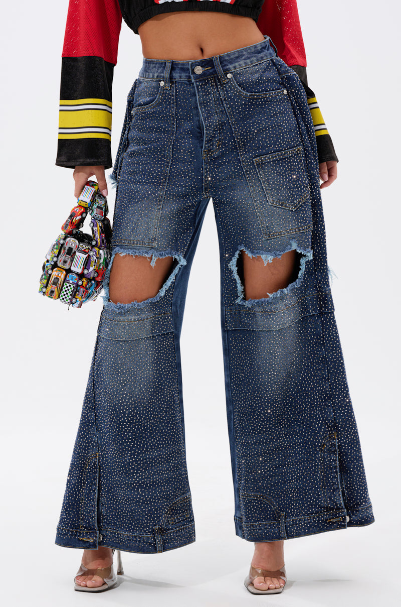 A person wearing SPARKLE QUEEN WIDE-LEG DENIM with rhinestones and large frayed knee rips, holding a colorful beaded handbag, and standing in clear high-heeled shoes.