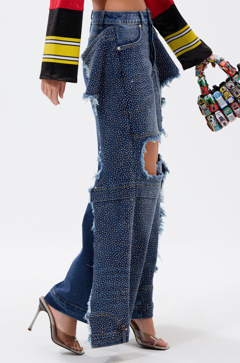 A woman styles the SPARKLE QUEEN WIDE-LEG DENIM—sparkly, high-waisted jeans with rhinestone details and a thigh rip—with a bold yellow striped long-sleeve top, heeled sandals, and a vibrant bag.