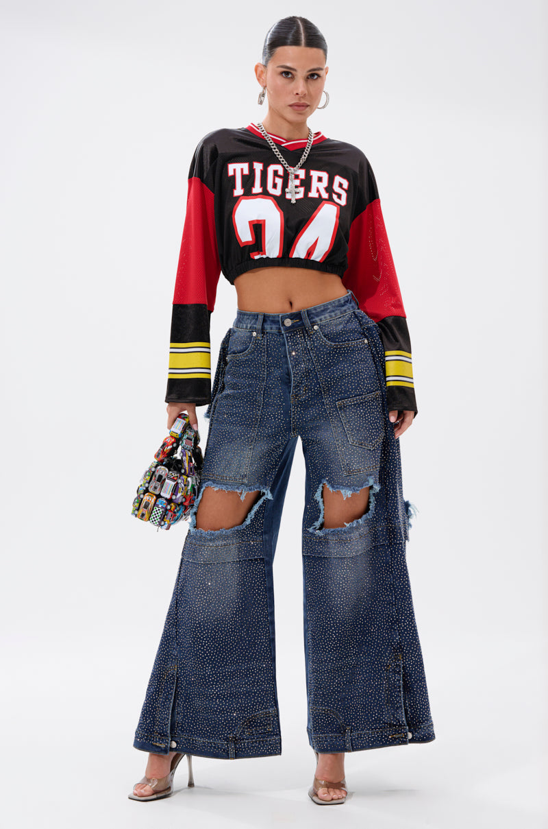 A woman poses against a white background wearing a cropped “Tigers 24” sports jersey, clear high heels, a colorful patterned handbag, and SPARKLE QUEEN WIDE-LEG DENIM featuring large ripped holes and sparkle details.