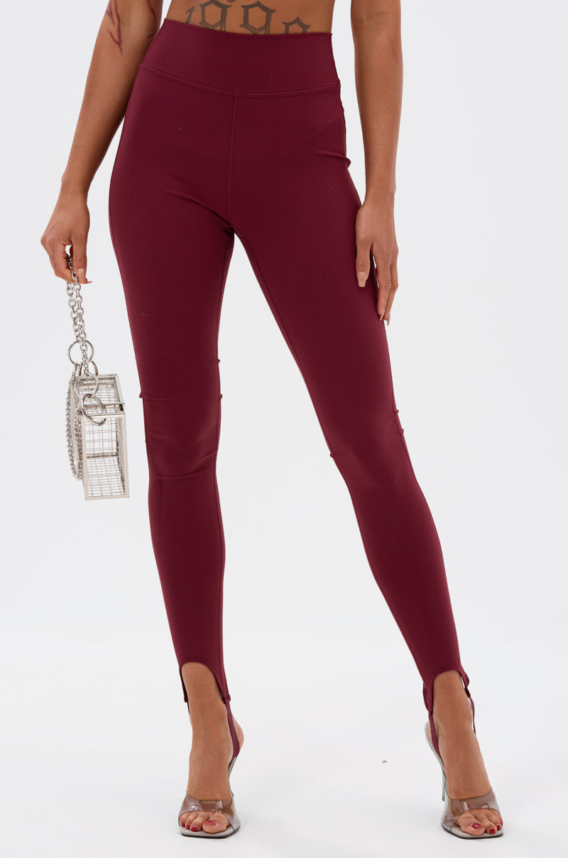 A person wears the DRIVE ME CRAZY STIRRUP LEGGING IN BURGUNDY, featuring a high-rise fit and front ankle slits, paired with clear high heels and a small silver chain handbag. The background is plain white.
