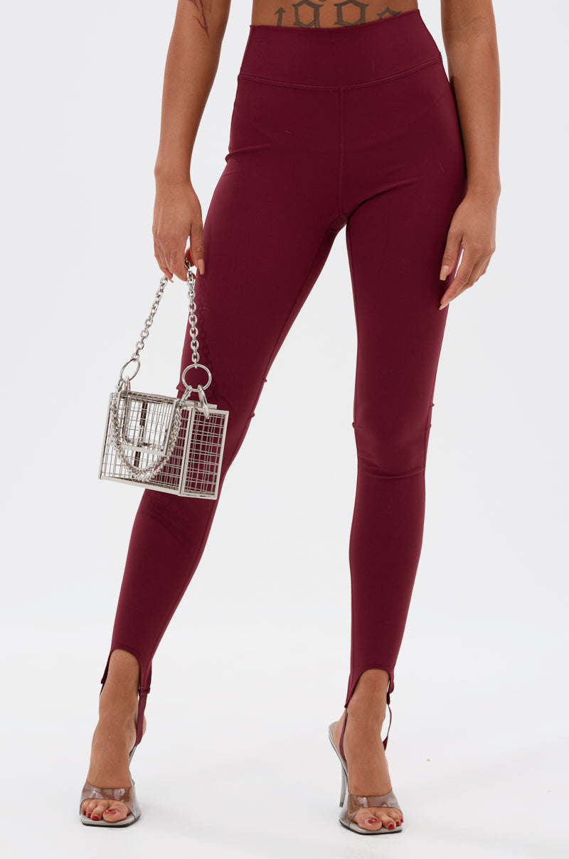 A woman wears the DRIVE ME CRAZY STIRRUP LEGGING IN BURGUNDY with front ankle slits, paired with clear high-heeled sandals and a small silver handbag, against a plain white background.