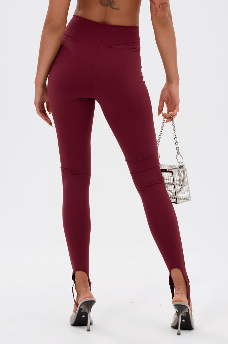 A person is shown from the back in DRIVE ME CRAZY STIRRUP LEGGING IN BURGUNDY, featuring high-rise fit and heel cutouts, styled with silver heels and a small metallic handbag against a plain white background.