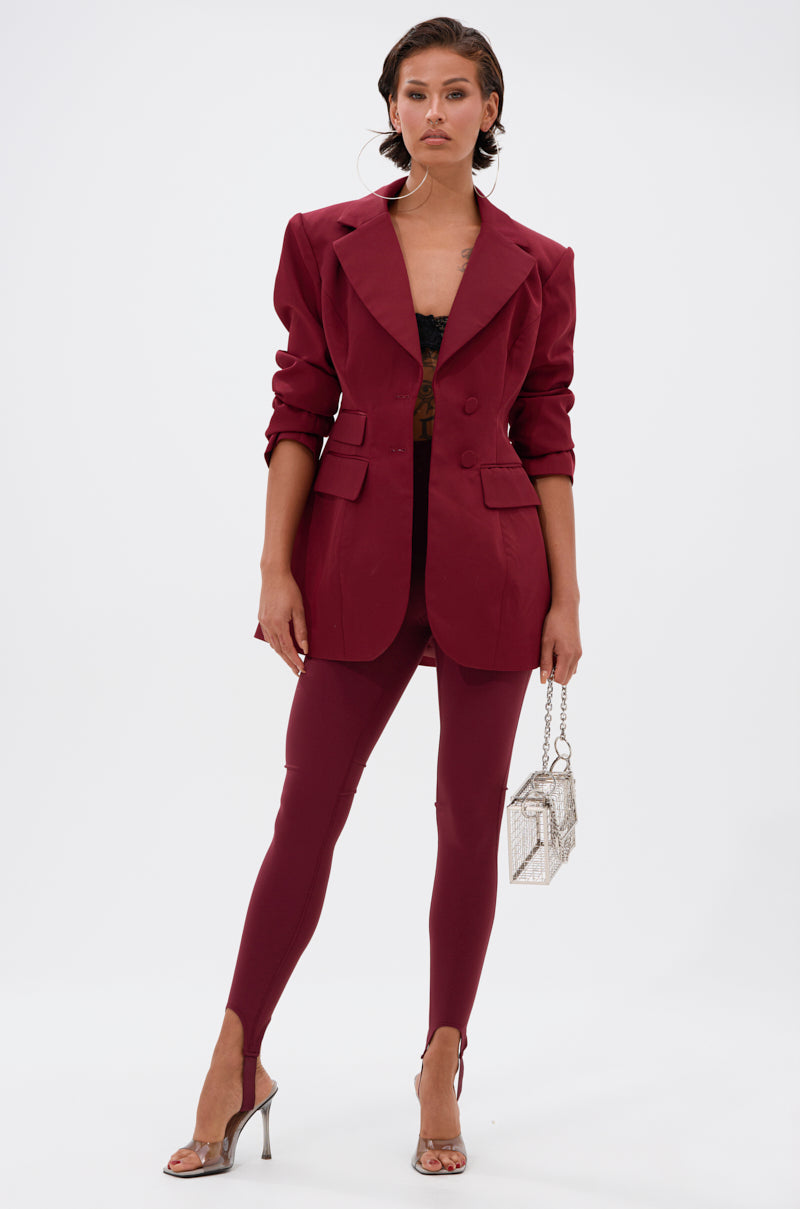 A woman poses in a stylish maroon pantsuit with a matching blazer, black lace top, silver high heels, and small silver handbag, featuring the DRIVE ME CRAZY STIRRUP LEGGING IN BURGUNDY, against a plain white background.