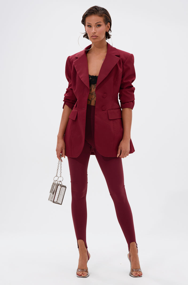 A woman poses confidently against a white background wearing the DRIVE ME CRAZY STIRRUP LEGGING IN BURGUNDY, a matching blazer, black lace bralette, clear heels, and holding a small silver chain handbag.