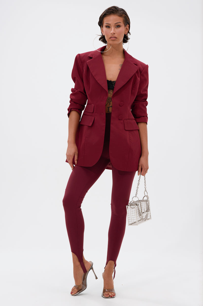 A woman models the DRIVE ME CRAZY STIRRUP LEGGING IN BURGUNDY with a matching jacket and black lace top, accessorized with a small silver handbag and clear high heels, her hair slicked back against a white background.