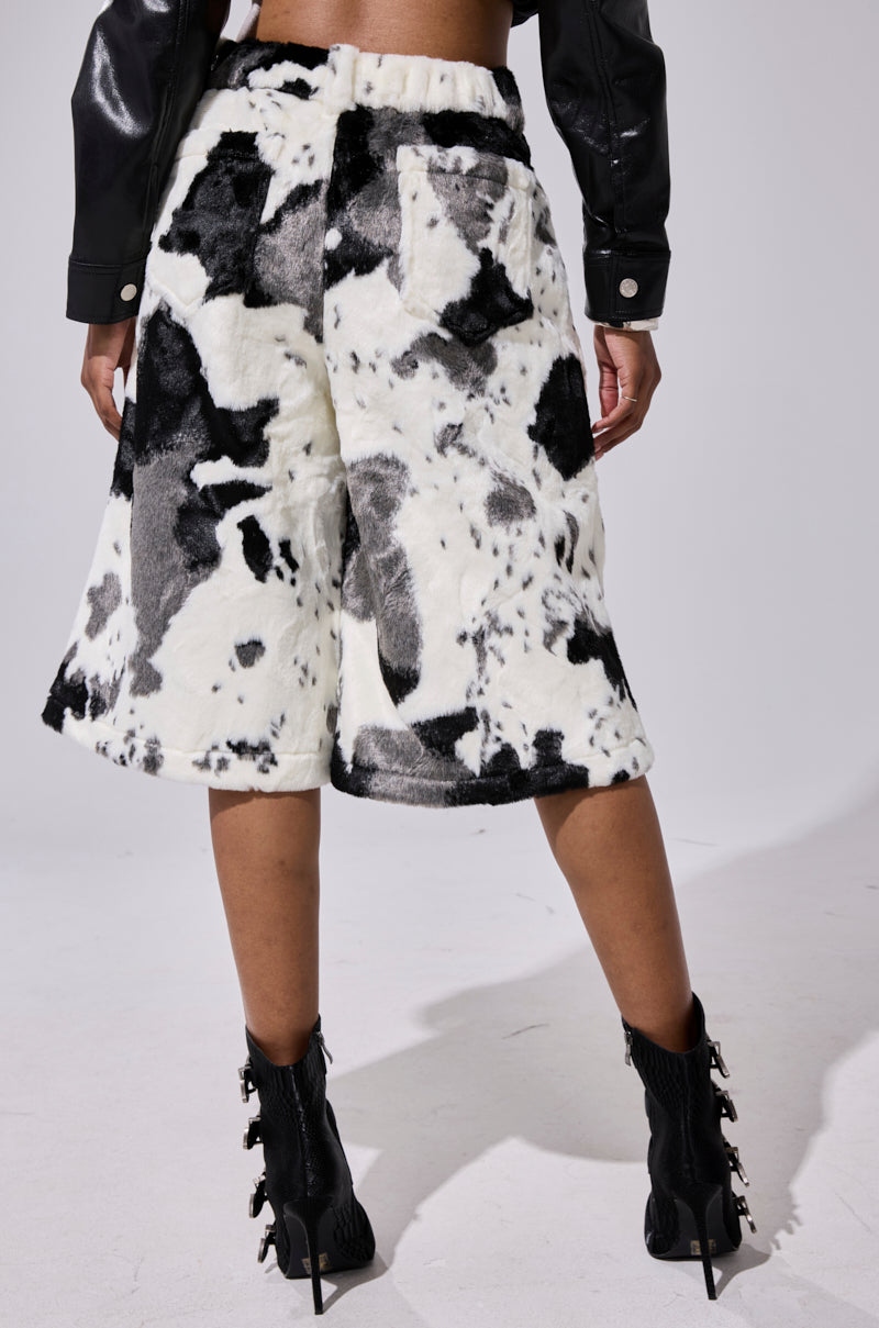 A person is seen from behind wearing the WATCH OUT COW BERMUDA SHORT, featuring a high-rise waistband, paired with black heeled boots and a black leather jacket against a plain light background. 3