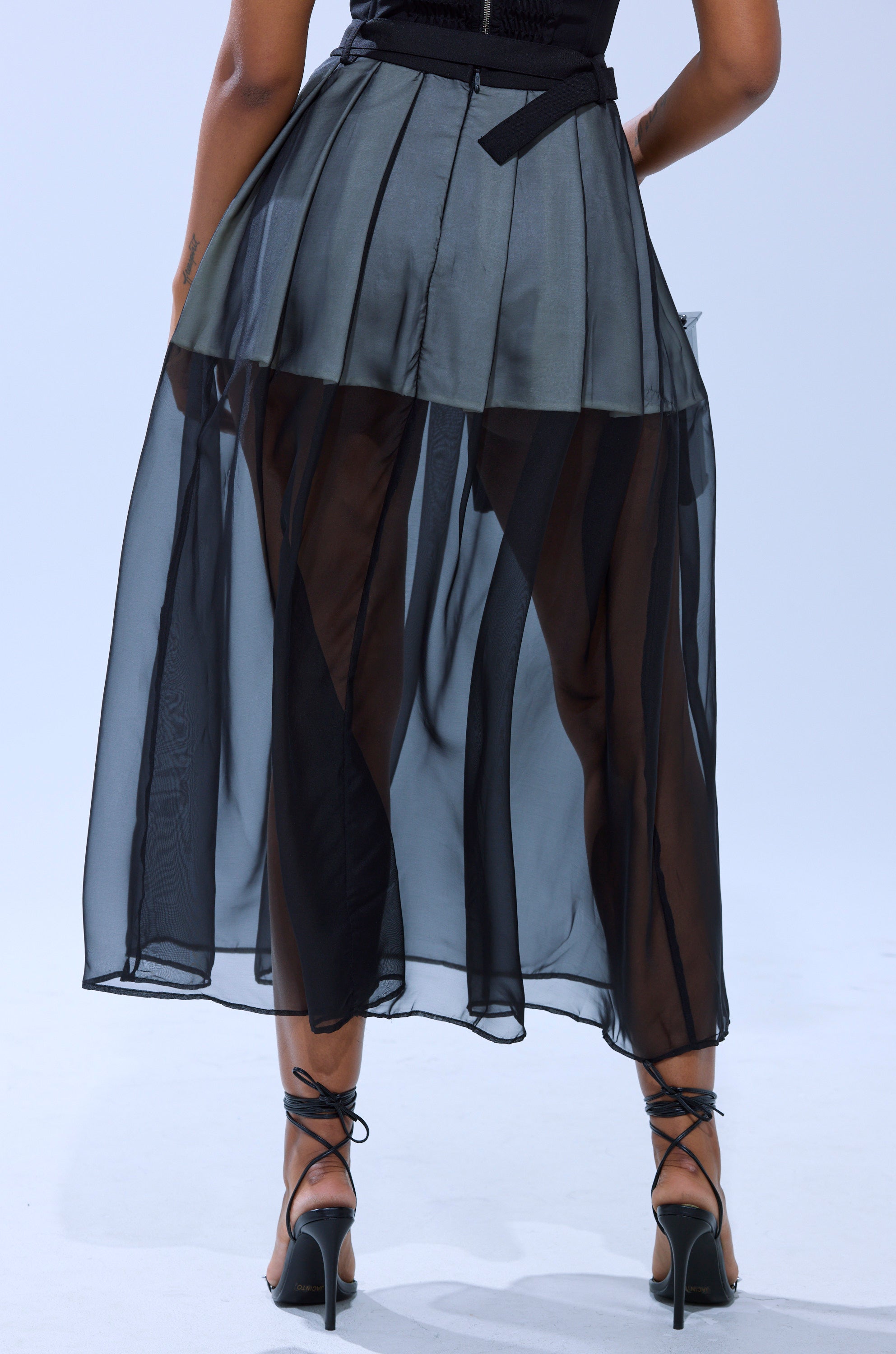 A person wearing the PARTY GIRL MIDI SKIRT featuring sheer mesh and vertical panels with strappy black high-heeled sandals, photographed from behind against a light background.