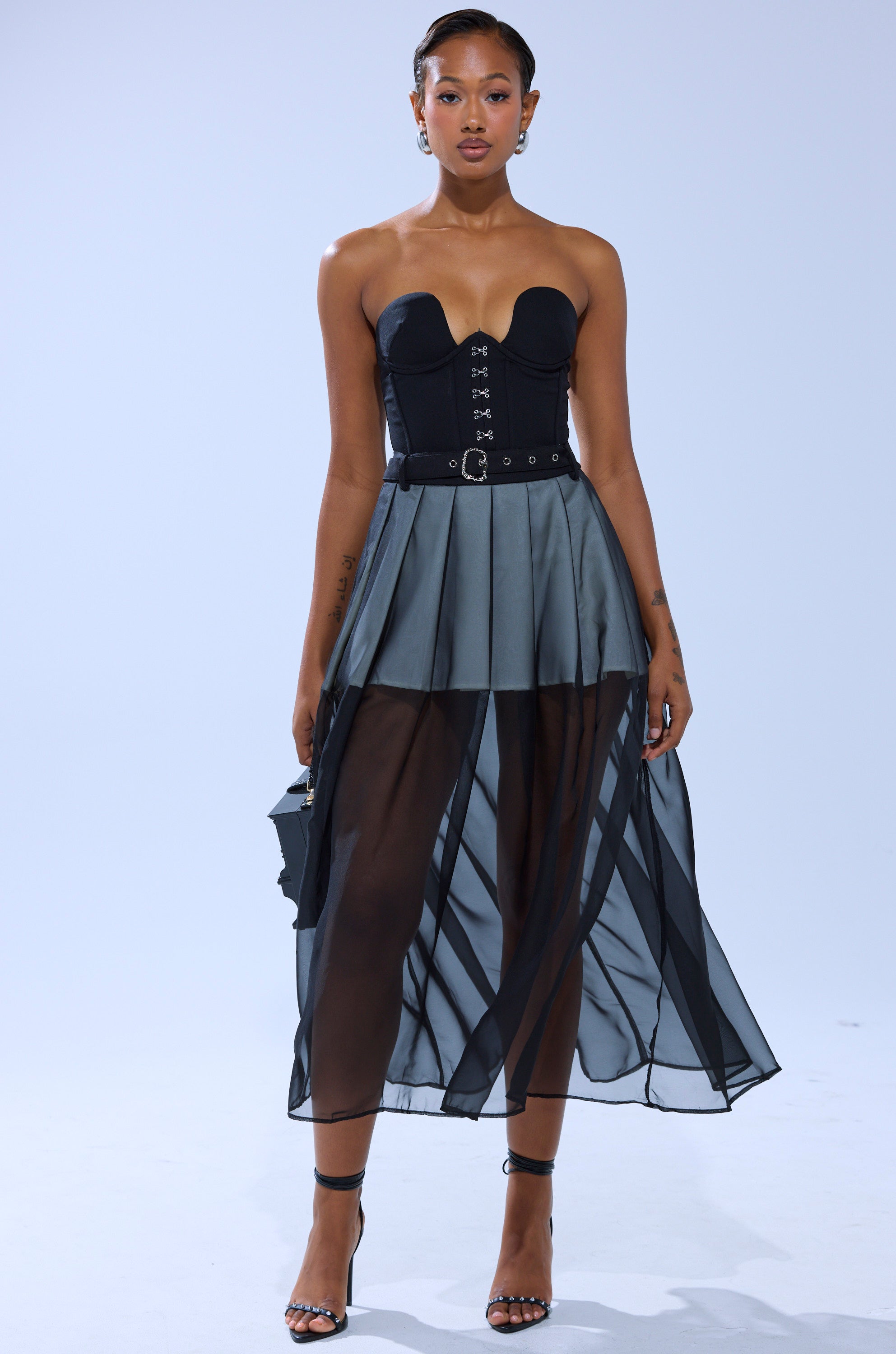 A woman stands against a plain background wearing the PARTY GIRL MIDI SKIRT, a strapless black corset top, strappy heels, and holding a small black handbag.