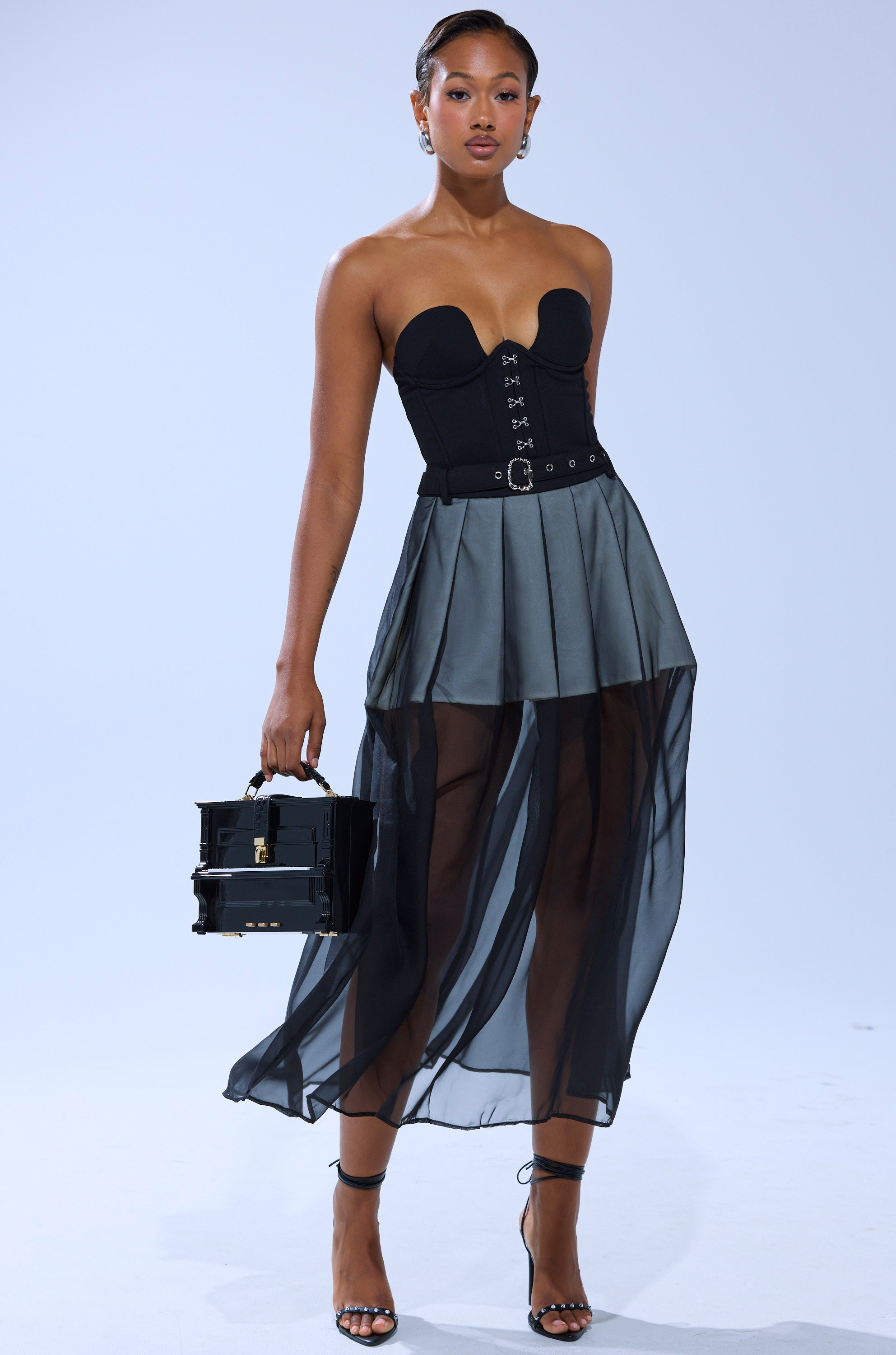 A woman models the PARTY GIRL MIDI SKIRT, a sheer mesh, high-waisted midi style. She pairs it with a black strapless corset top, strappy heels, a black handbag, and wears her hair sleeked back against a plain light background.