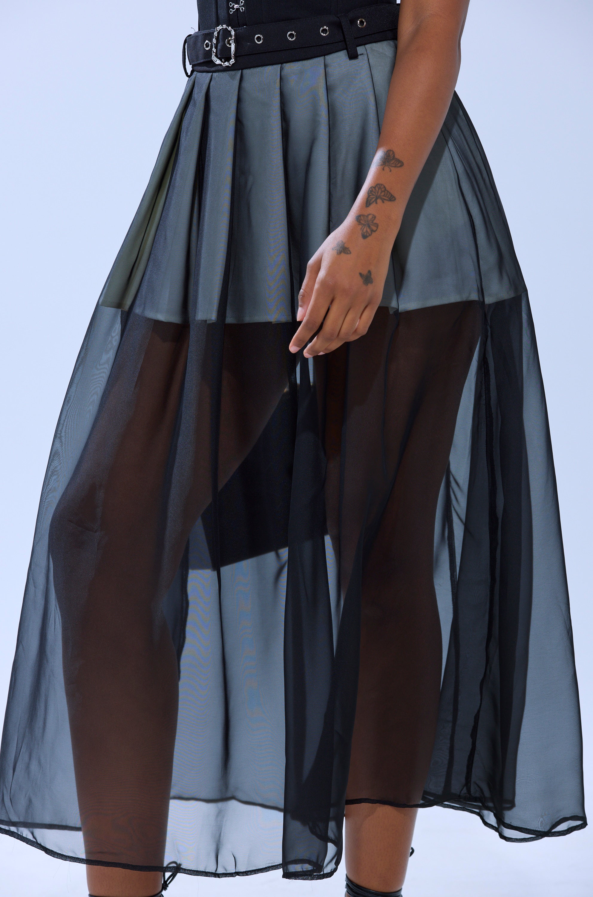 A person wears the PARTY GIRL MIDI SKIRT, a black sheer mesh pleated design over black shorts with a black belt, showcasing hand and forearm tattoos against a plain light background.