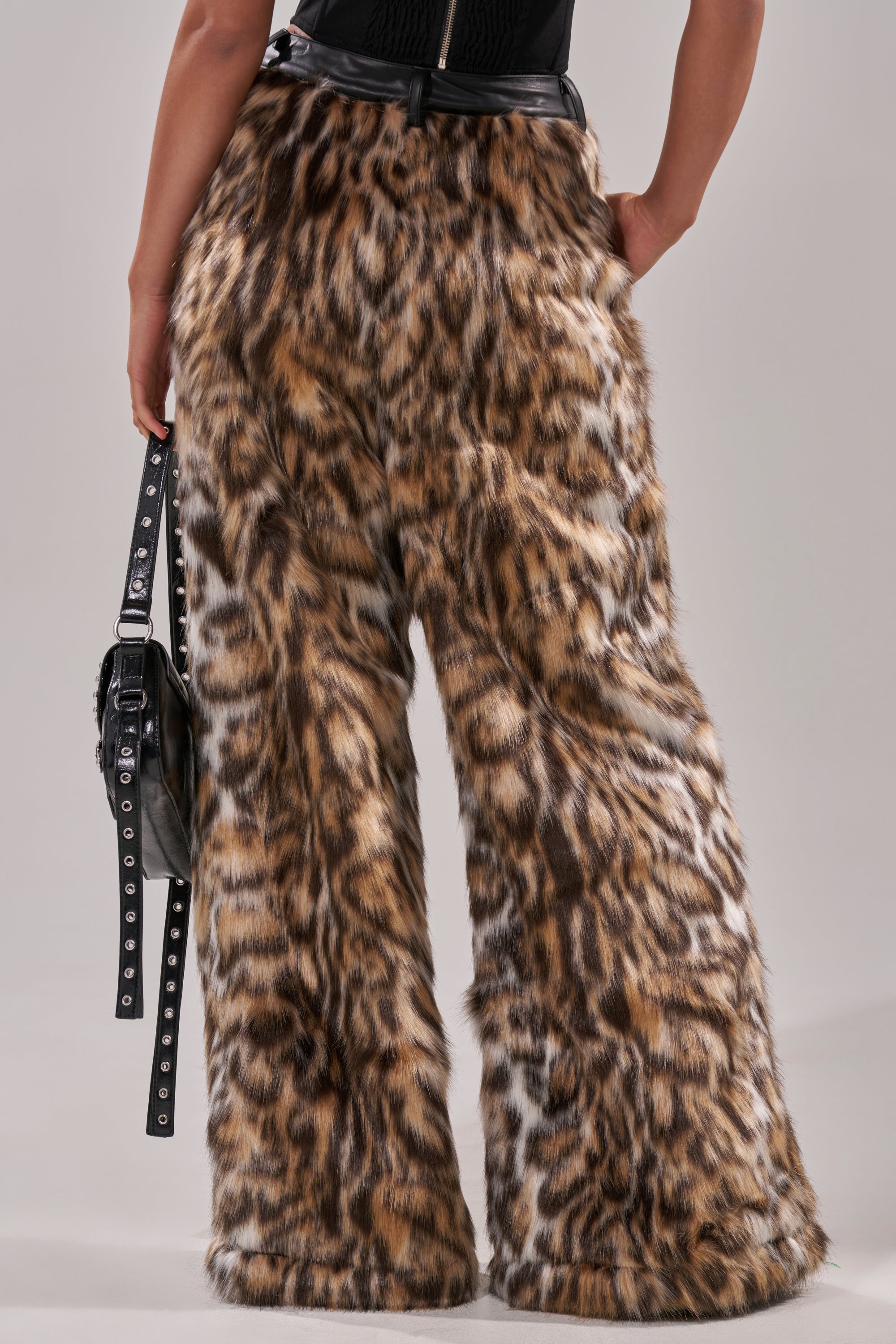 A person is seen from the back wearing CATCH ME IF YOU CAN LEOPARD FUR TROUSER—wide-leg, high-rise faux fur pants with a bold leopard print—styled with a black belt and holding a black studded handbag.