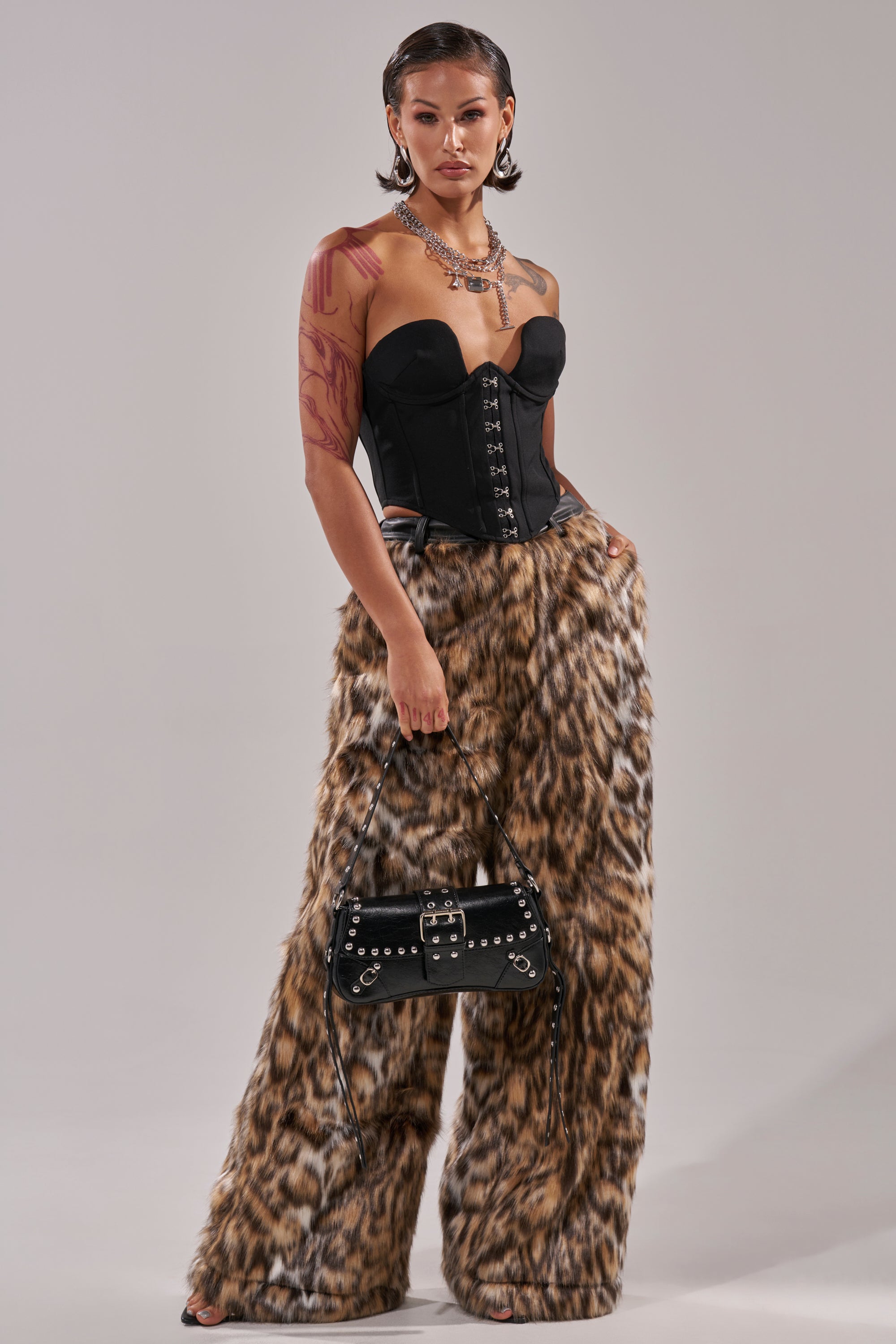 A woman exudes confidence in the CATCH ME IF YOU CAN LEOPARD FUR TROUSER and a black strapless corset, accessorized with a studded black handbag, bold jewelry, slicked-back hair, and visible arm tattoos for an edgy finish.