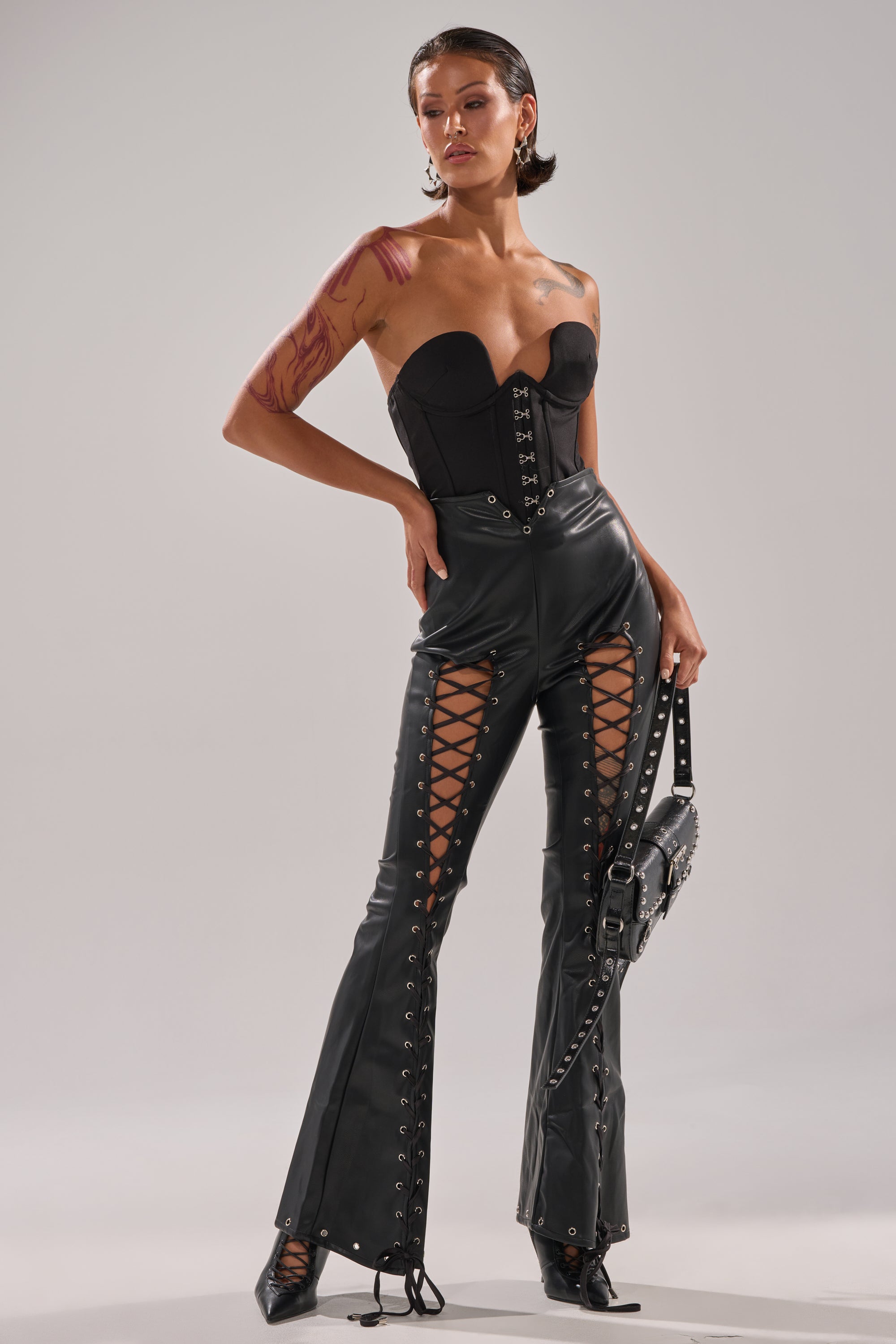 A woman poses confidently in the I KNOW THATS RIGHT FLARE FAUX LEATHER PANT with a black strapless corset top, studded handbag, and pointed boots; her slicked-back hair and visible arm tattoos complete her bold, edgy look.