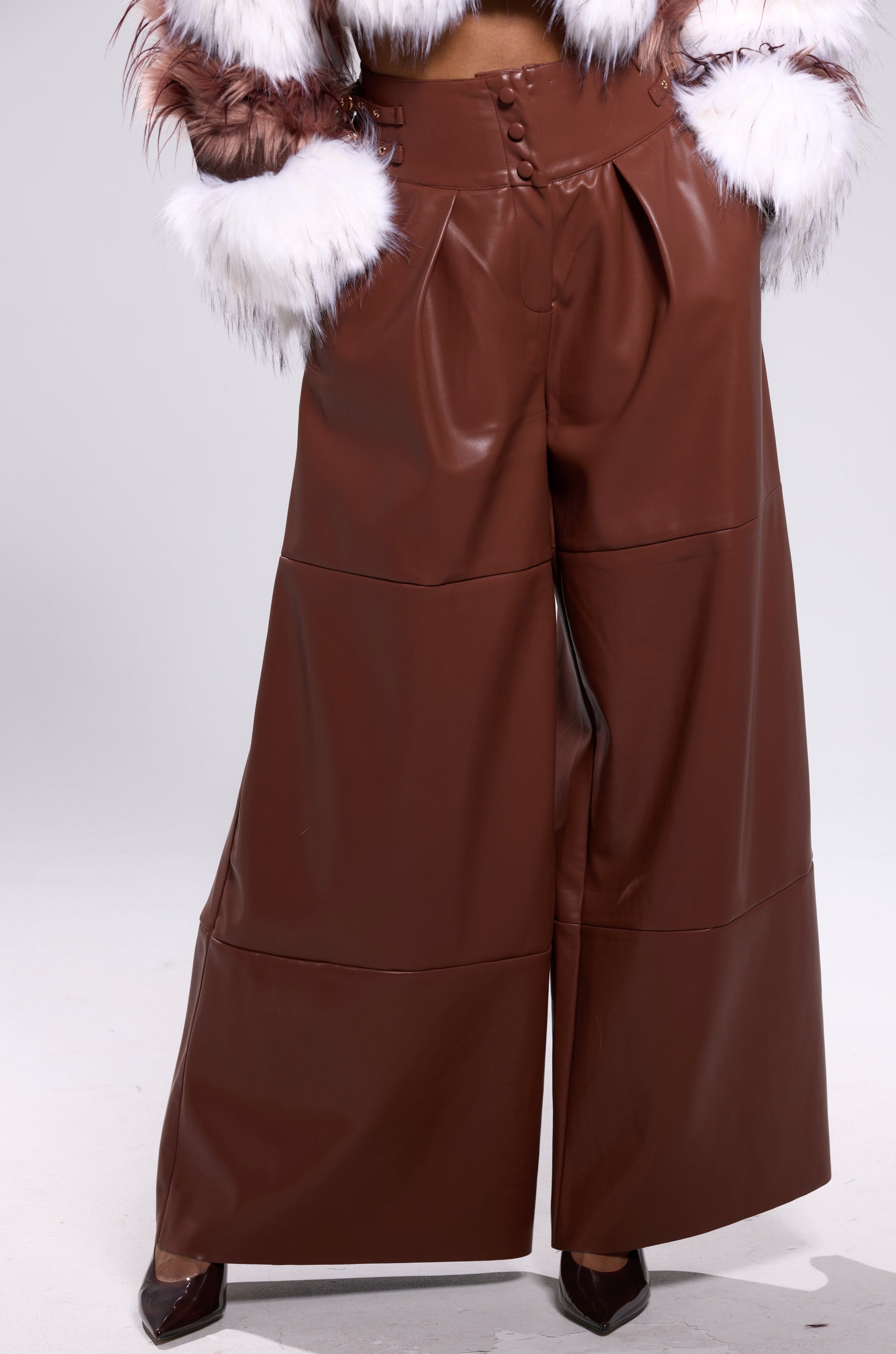A person stands against a plain light background wearing BETTER OFF WITHOUT YOU FAUX LEATHER TROUSER, a jacket with white and brown fur cuffs, and pointed brown shoes.