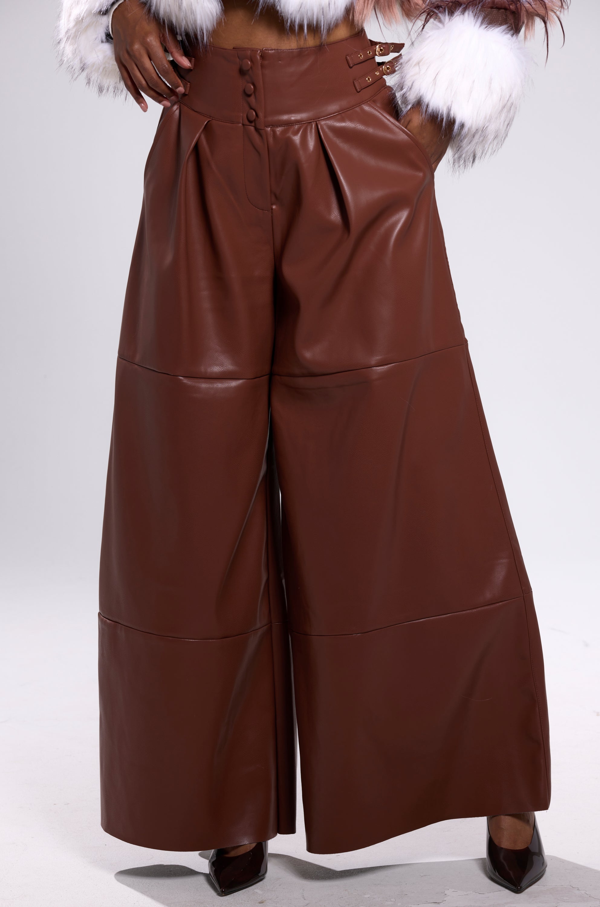 A person wears the BETTER OFF WITHOUT YOU FAUX LEATHER TROUSER in brown, featuring a high-rise fit, wide legs, buttons, and pockets. The visible part of the outfit is only the lower half.