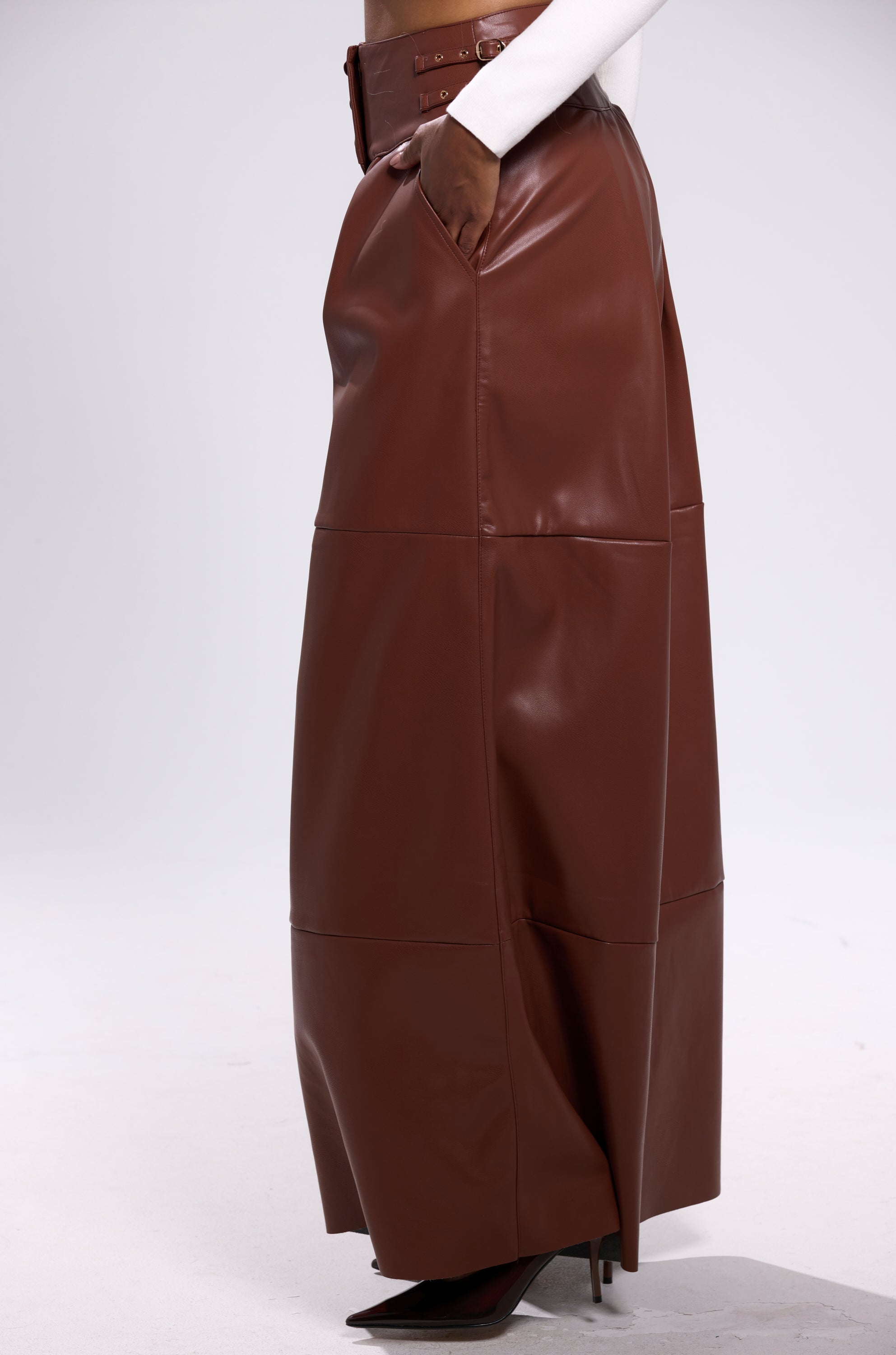 A person wears the BETTER OFF WITHOUT YOU FAUX LEATHER TROUSER in brown, styled with a white top and pointed-toe heeled boots. Their hand is in the pocket, set against a plain light gray background.