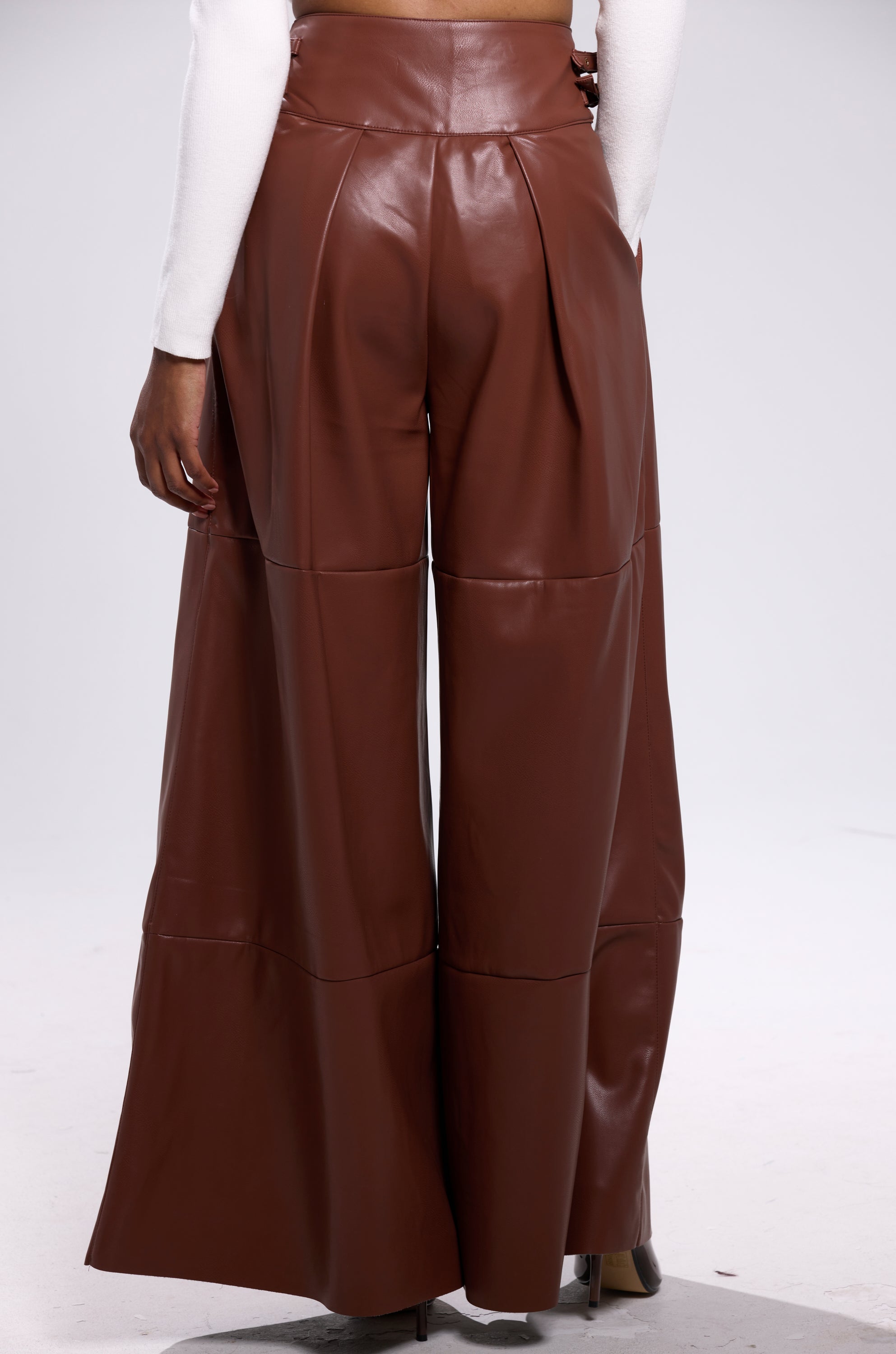 Someone stands facing away from the camera against a plain light background, wearing BETTER OFF WITHOUT YOU FAUX LEATHER TROUSER in brown with a high-rise fit and wide legs, paired with a long-sleeve white top.
