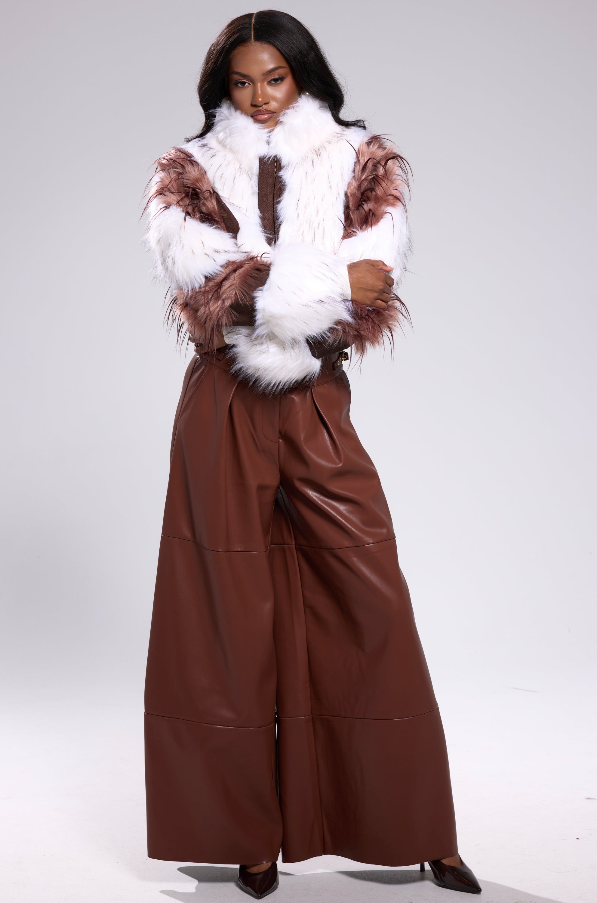 A woman with long dark hair poses confidently against a light background, wearing the BETTER OFF WITHOUT YOU FAUX LEATHER TROUSER in a wide-leg high-rise fit and a white faux fur jacket with brown accents, arms crossed.