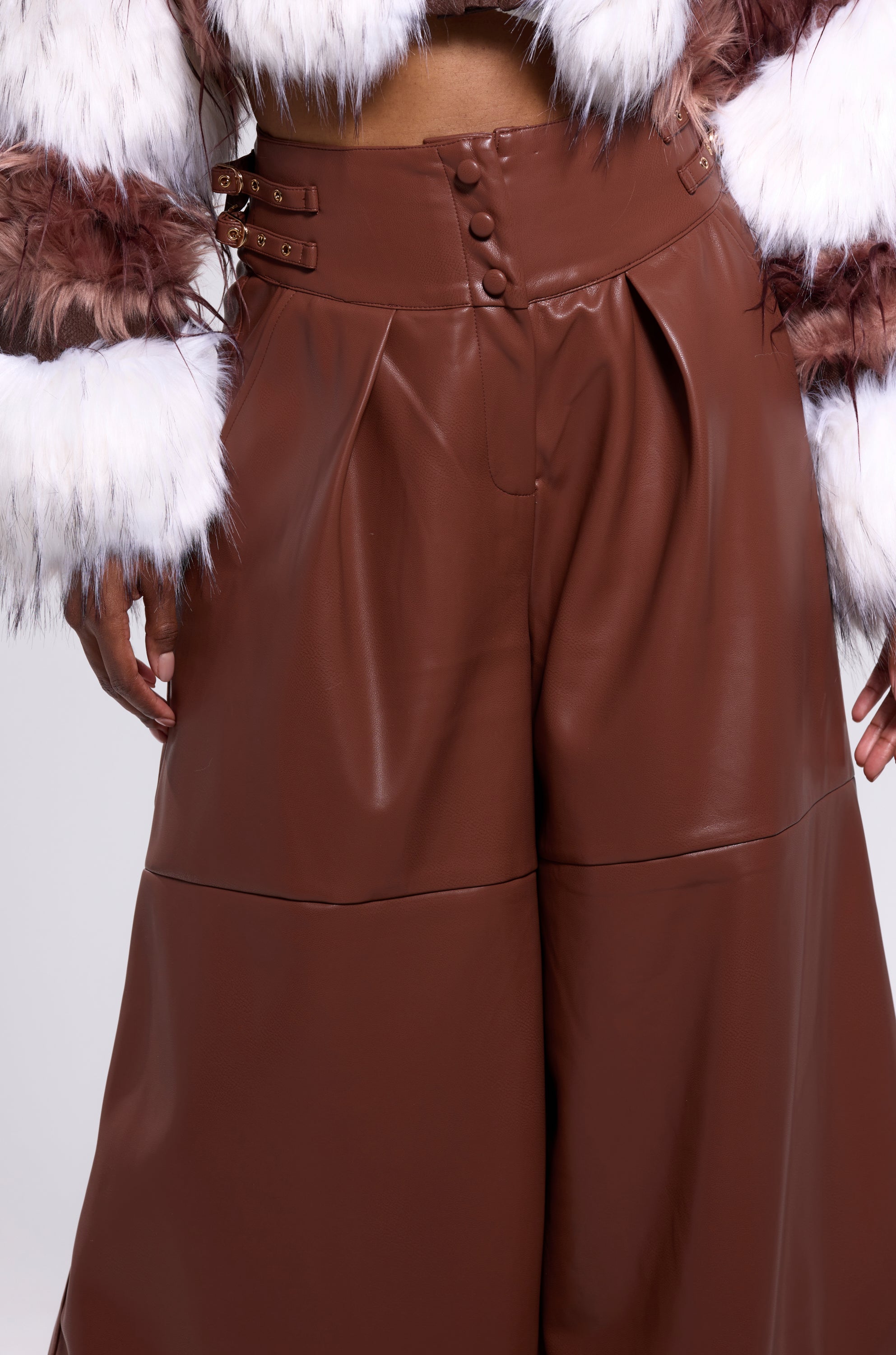Someone wears the BETTER OFF WITHOUT YOU FAUX LEATHER TROUSER, featuring a high-rise wide-leg cut and belt buckle details, styled with a cropped brown and white fluffy fur jacket. The photo is cropped from chest to knees.