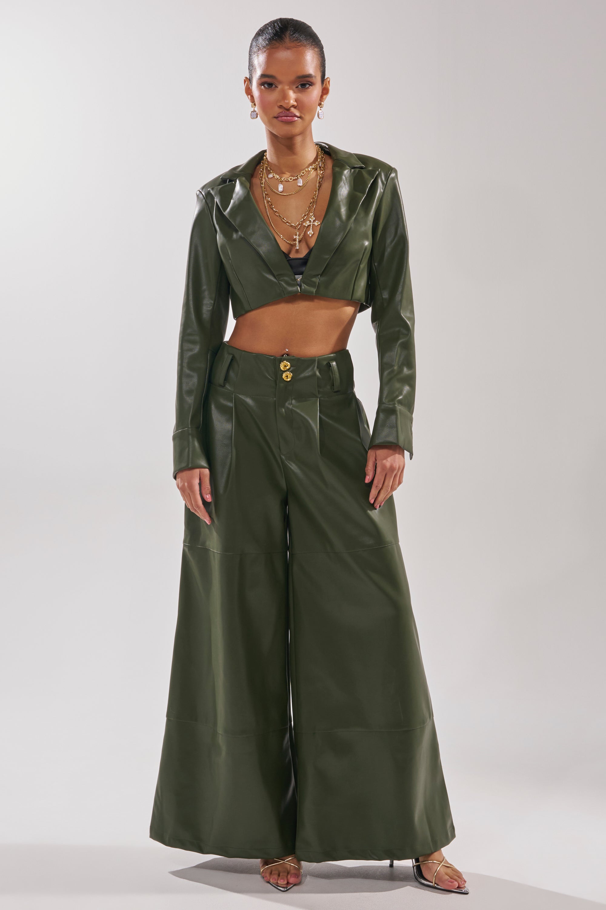 A woman stands confidently in REMEMBER YOU WIDE LEG TROUSER and a matching olive green faux leather cropped jacket, styled with layered necklaces and strappy heels against a plain light background. 0