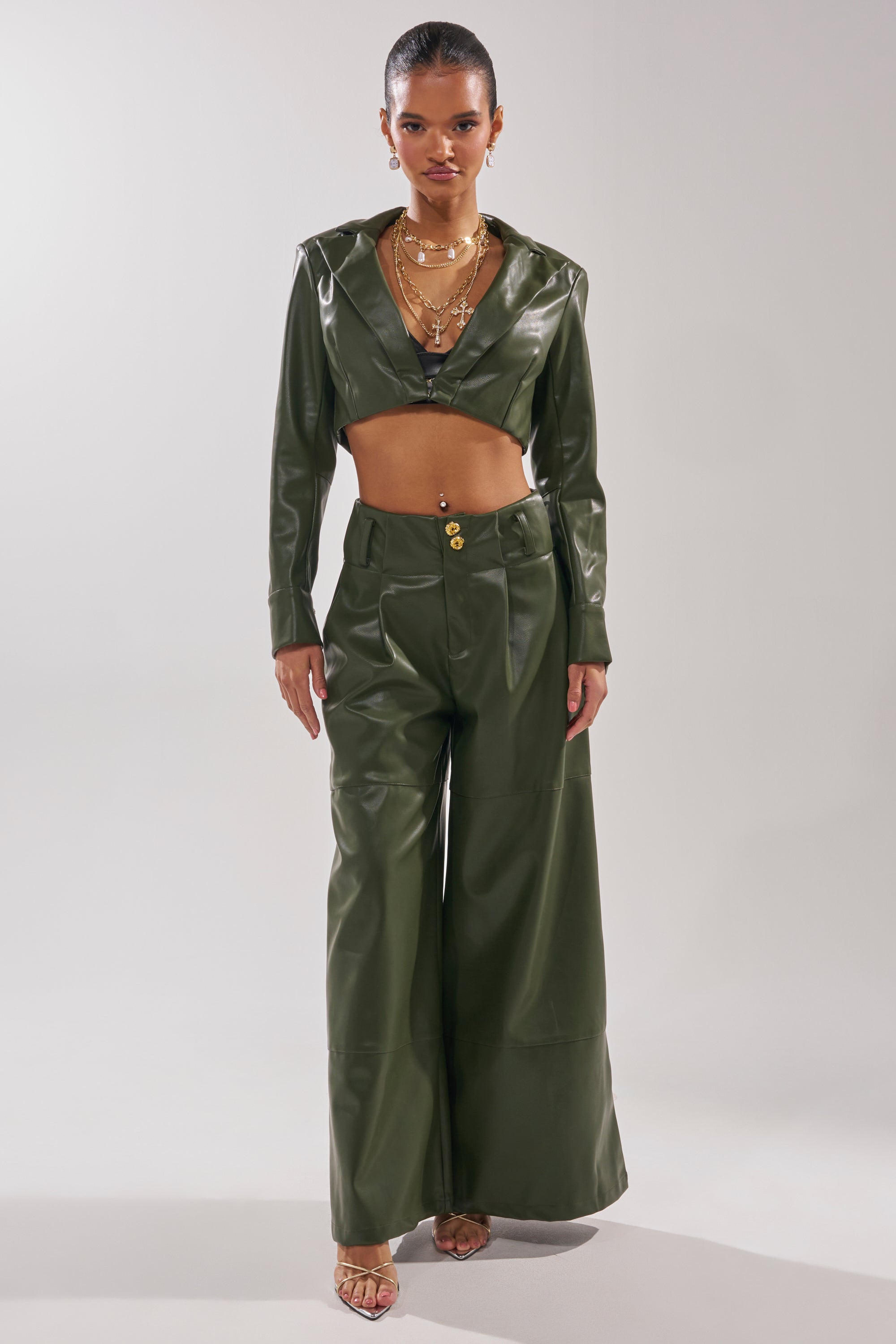 A woman faces forward, wearing the REMEMBER YOU WIDE LEG TROUSER in shiny olive green with a cropped, long-sleeved jacket. Her hair is tied back, and she accessorizes with layered necklaces and open-toe heels. 6