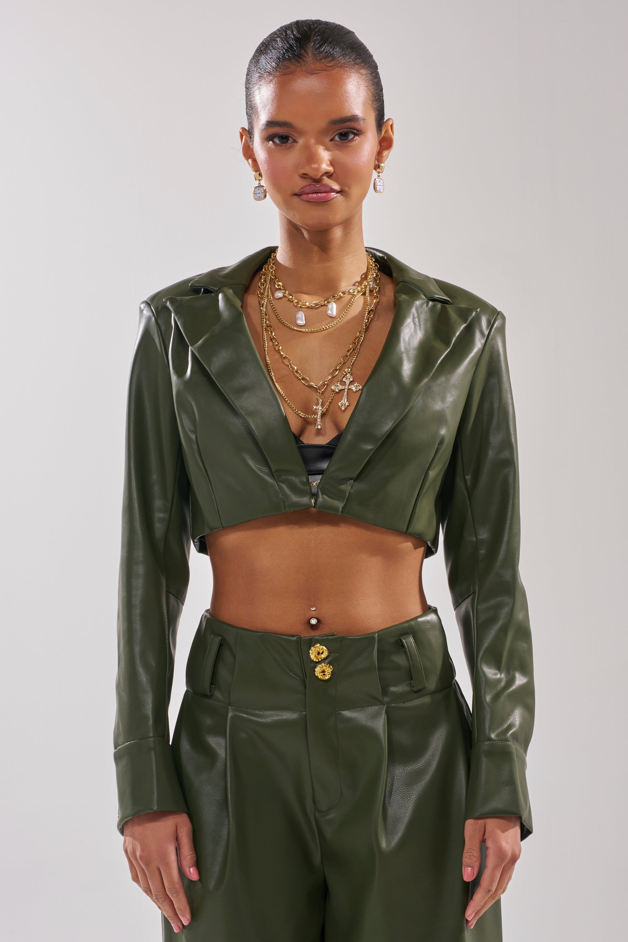 A woman models the REMEMBER YOU CROPPED BLAZER in shiny olive green faux leather with gold button cuffs, paired with matching high-waisted pants and layered gold jewelry, standing against a plain background. 1