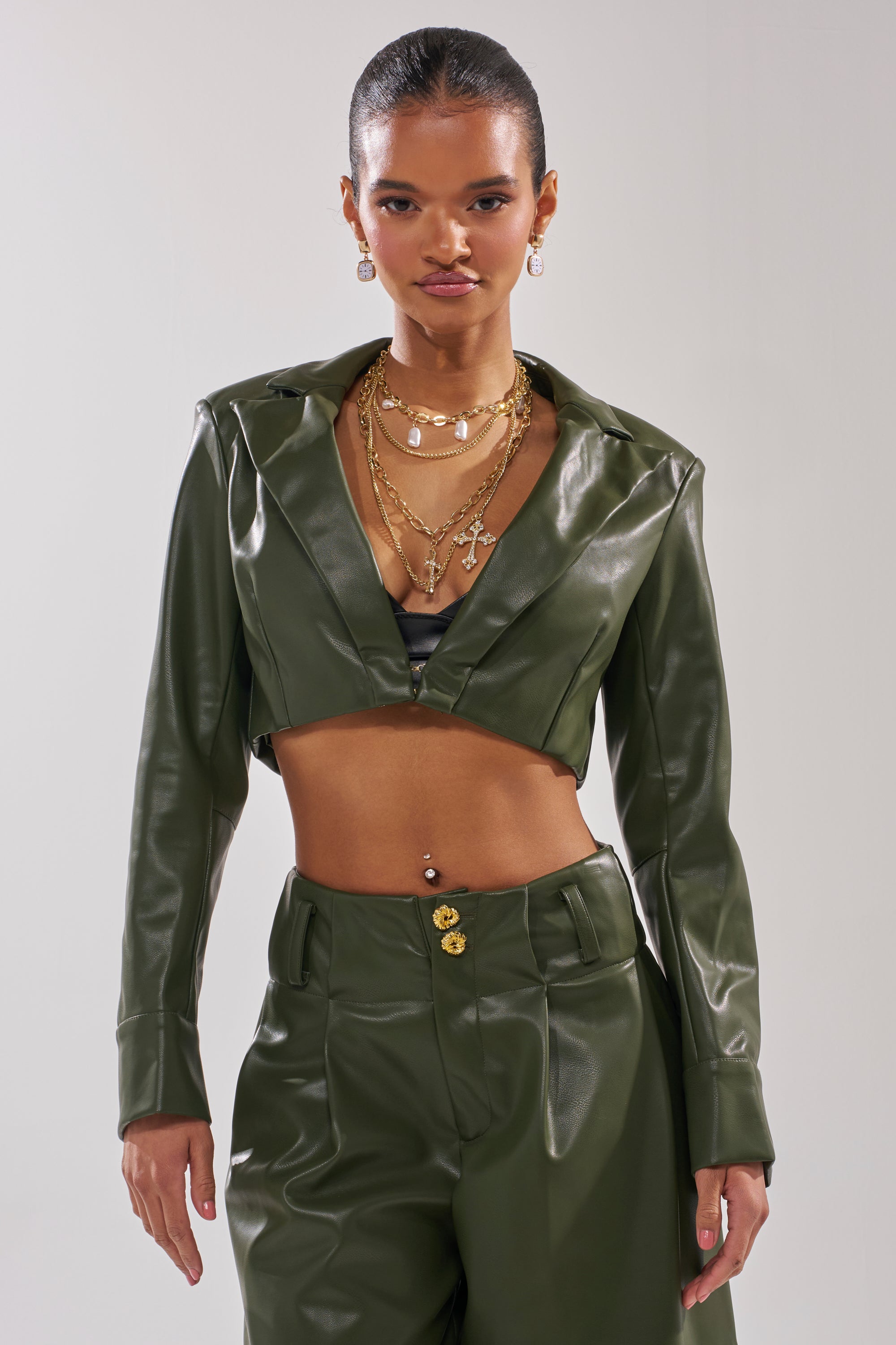A woman confidently stands against a plain background in the AKIRA Label REMEMBER YOU CROPPED BLAZER and matching green high-waisted pants, accessorized with layered gold necklaces, earrings, and a visible navel piercing. 2