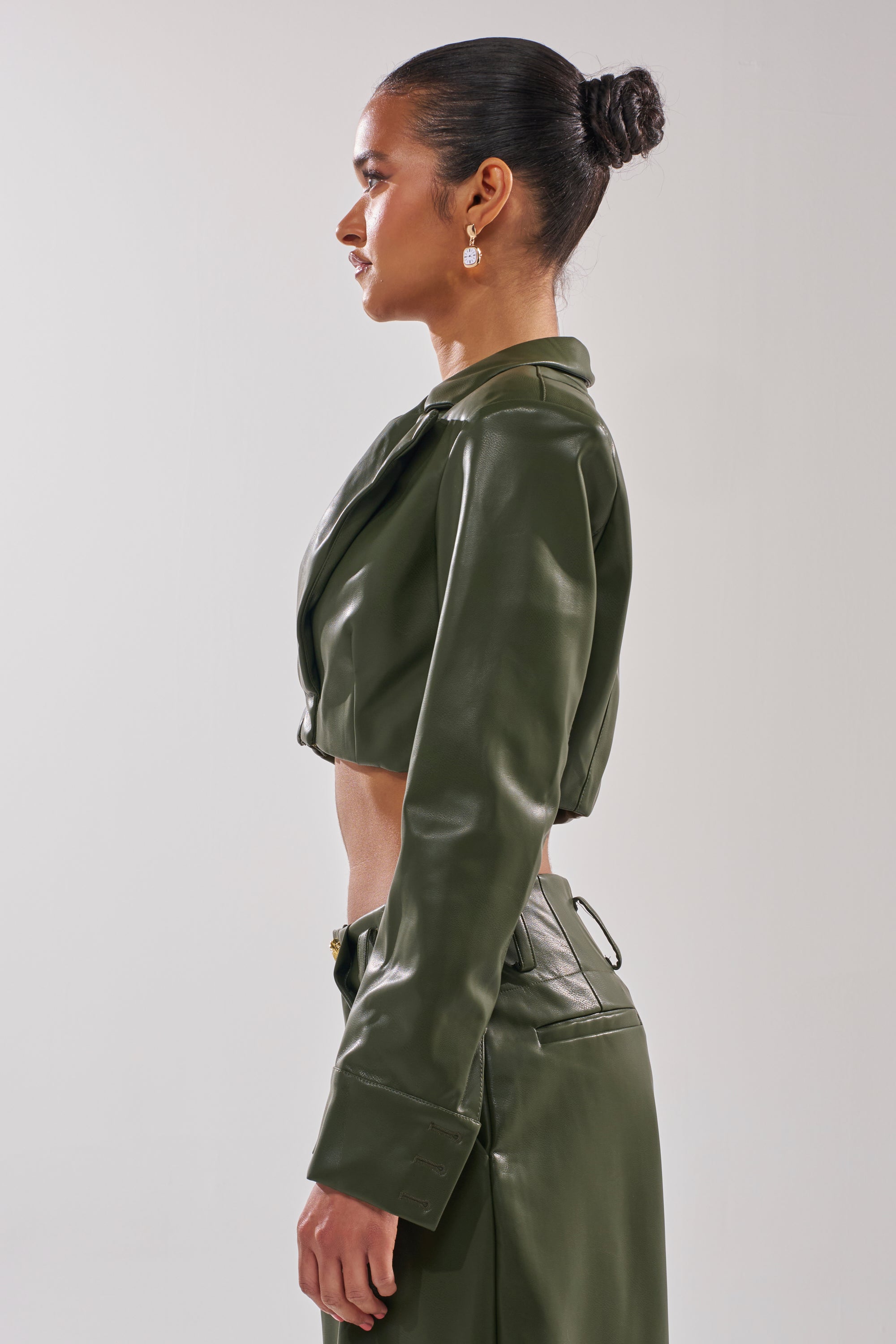 A woman with dark hair in a bun, gold earrings, and an olive green REMEMBER YOU CROPPED BLAZER with matching high-waisted pants stands in profile against a plain, light background. 3