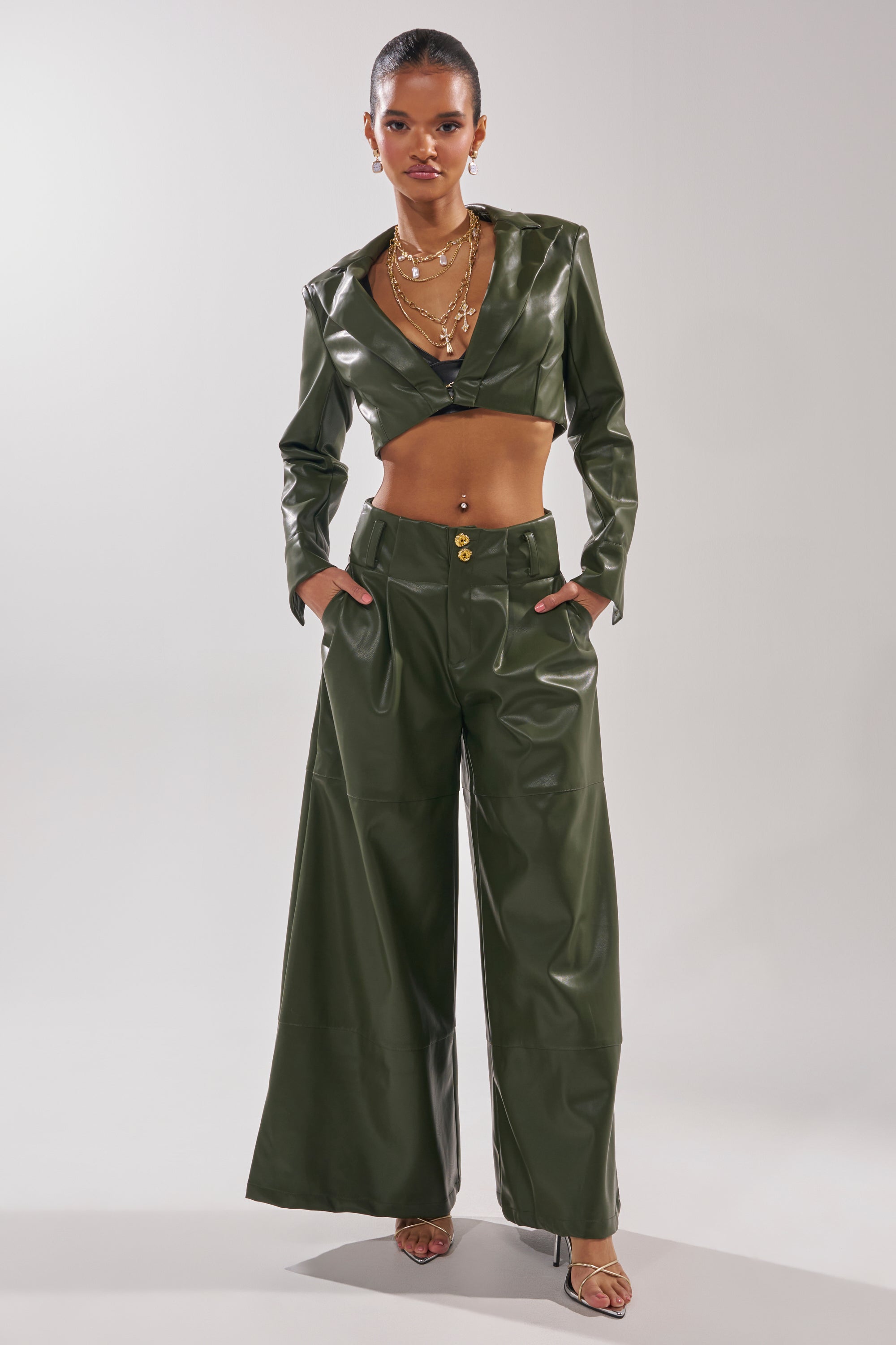 A woman stands confidently in the REMEMBER YOU CROPPED BLAZER, a shiny dark green faux leather piece paired with matching wide-leg pants, layered gold necklaces, and open-toe heels against a plain background. 6
