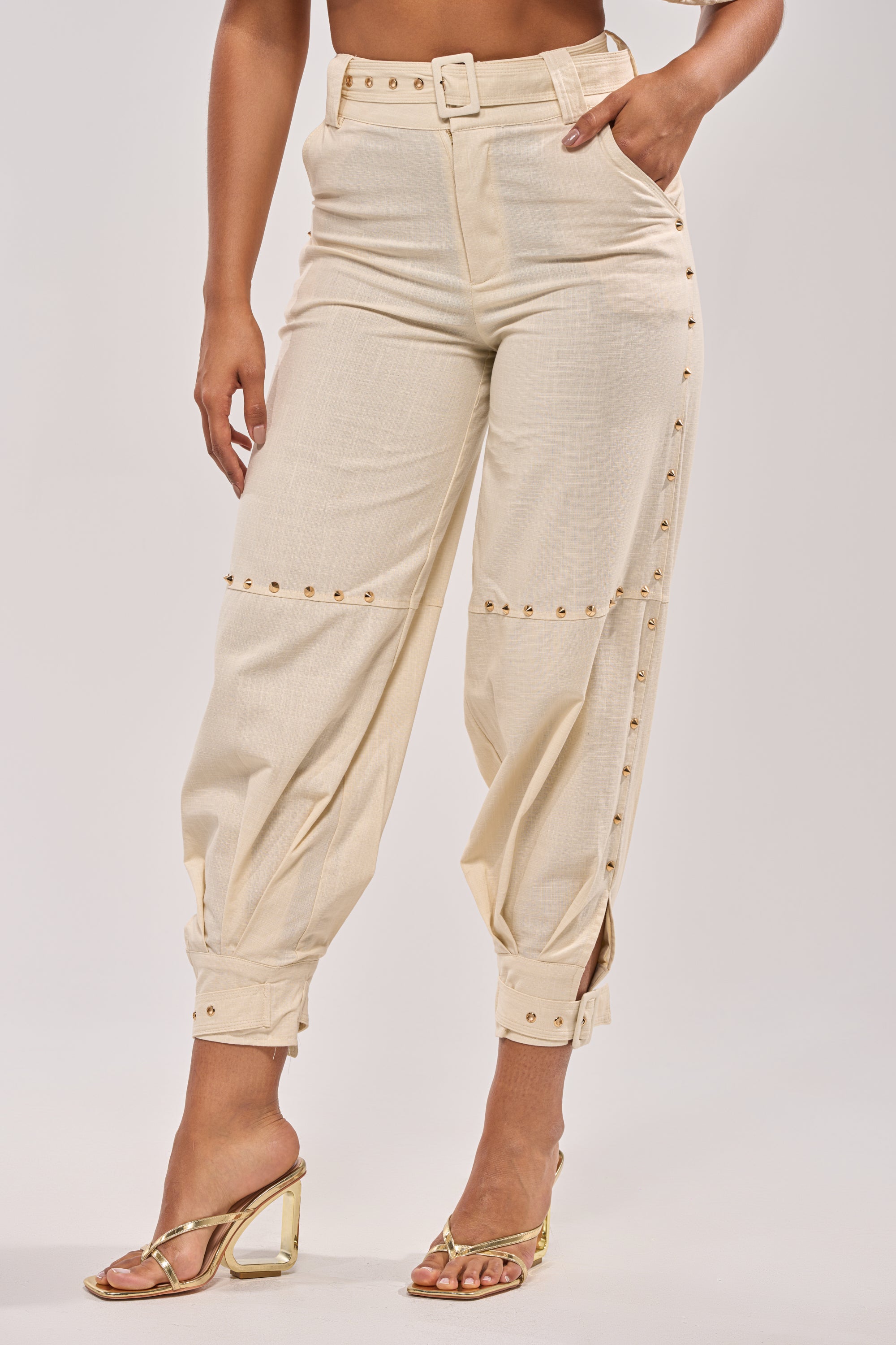 Wearing MAEVE LINEN STUDDED PANT—high-rise, belted, relaxed-fit pants cuffed at the ankles with gold side studs—paired with gold high-heeled sandals. Only the lower torso and legs are visible. 2